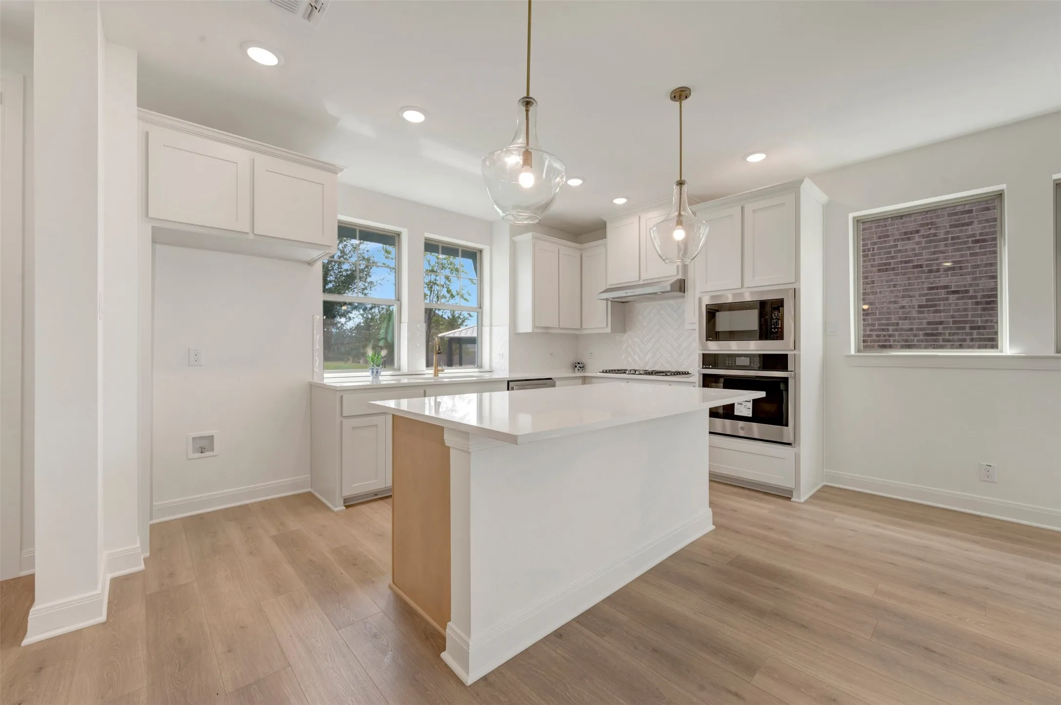 Modern kitchen featuring a central island with a light-toned countertop, light-finish flooring, white cabinetry, integrated stainless steel appliances, and two clear glass pendant lights