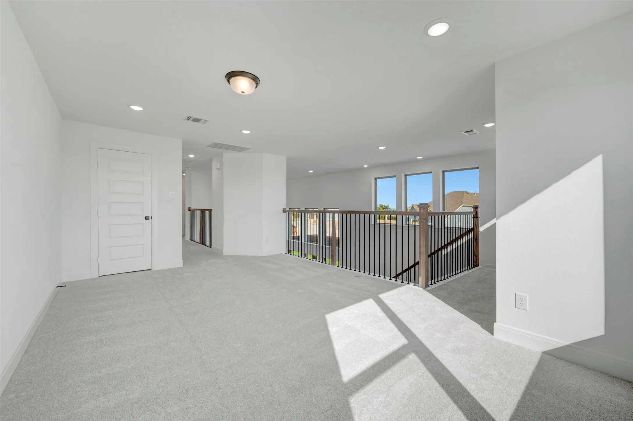 Carpeted upper-level landing featuring a wood and metal railing, three windows, a white five-panel door, and recessed lighting