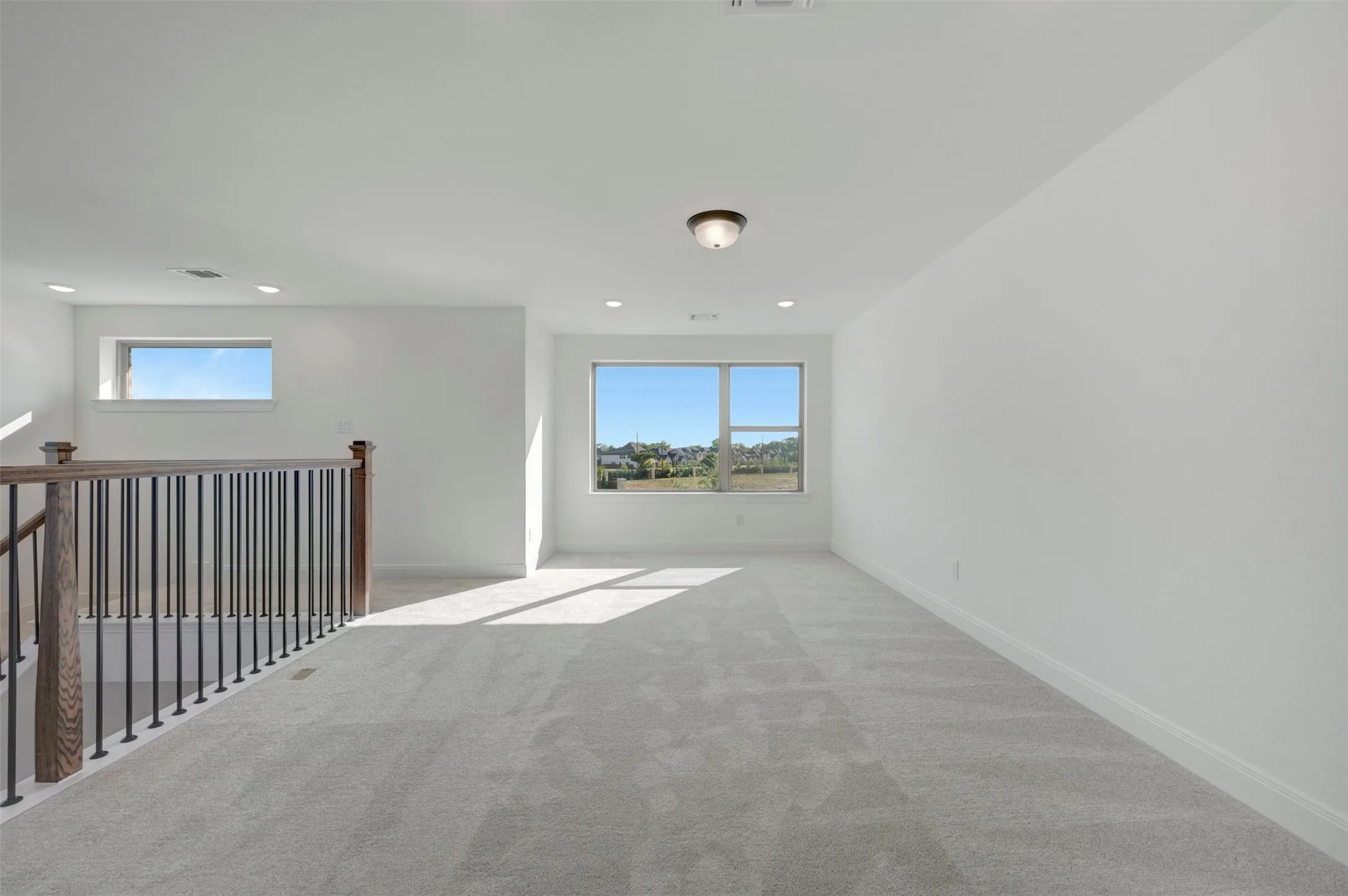 Spacious carpeted room featuring multiple windows, a wood and iron railing, a flush mount ceiling light, and recessed lighting