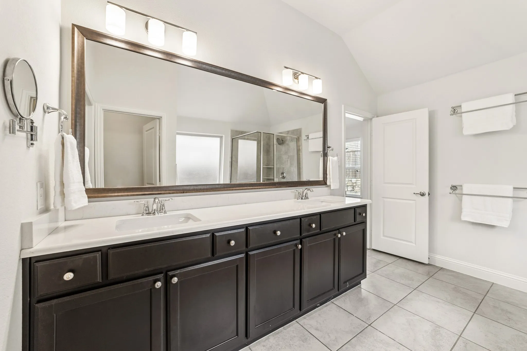 Full bathroom featuring a shower stall, double vanity, light tile patterned flooring, and vaulted ceiling