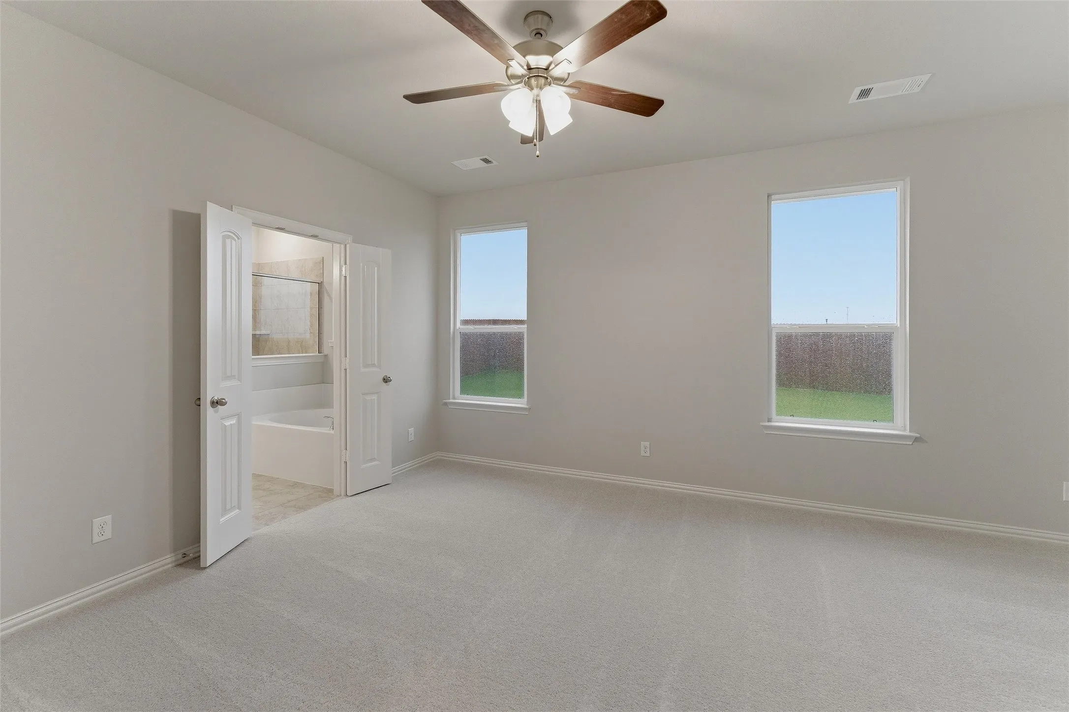 Spacious room featuring light gray carpeting, two windows with white trim, and a ceiling fan with wood-finish blades