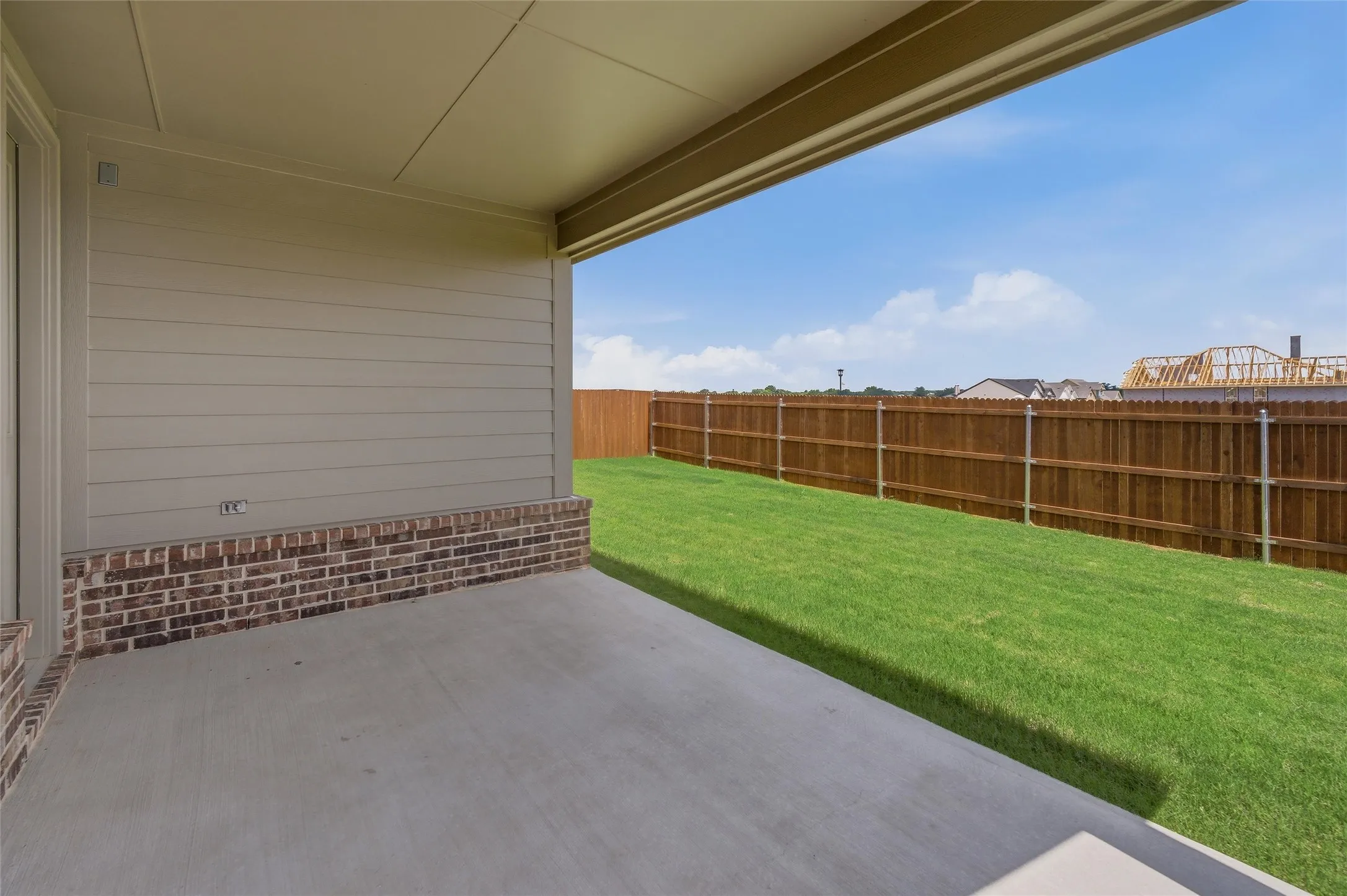 Covered concrete patio with a painted ceiling, brick and siding exterior, expansive green lawn, and a perimeter wood fence