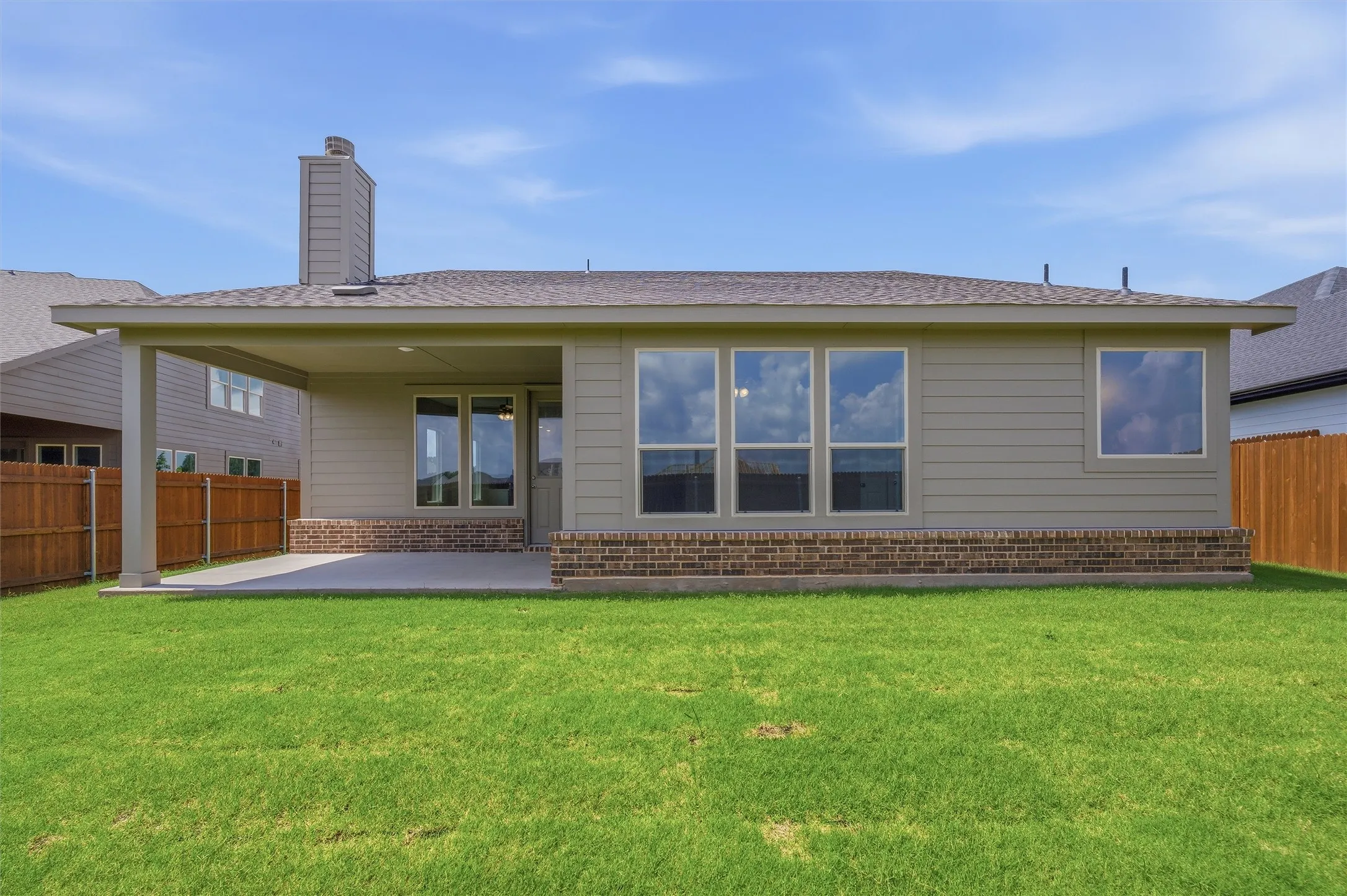 Expansive green lawn bordering a single-story home featuring a covered patio, brick foundation, and multiple windows