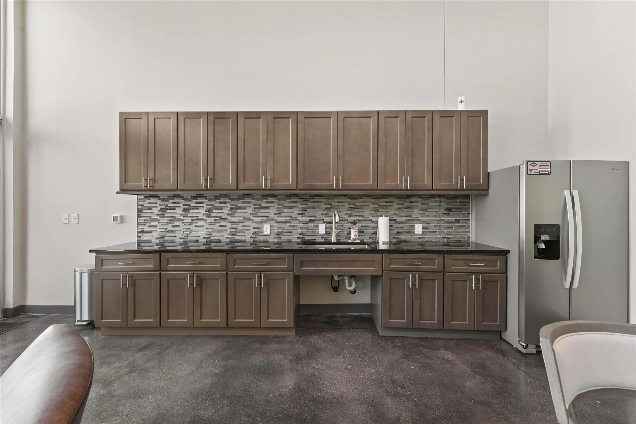 Kitchen featuring tasteful backsplash, stainless steel refrigerator with ice dispenser, dark brown cabinets, concrete flooring, and dark stone counters