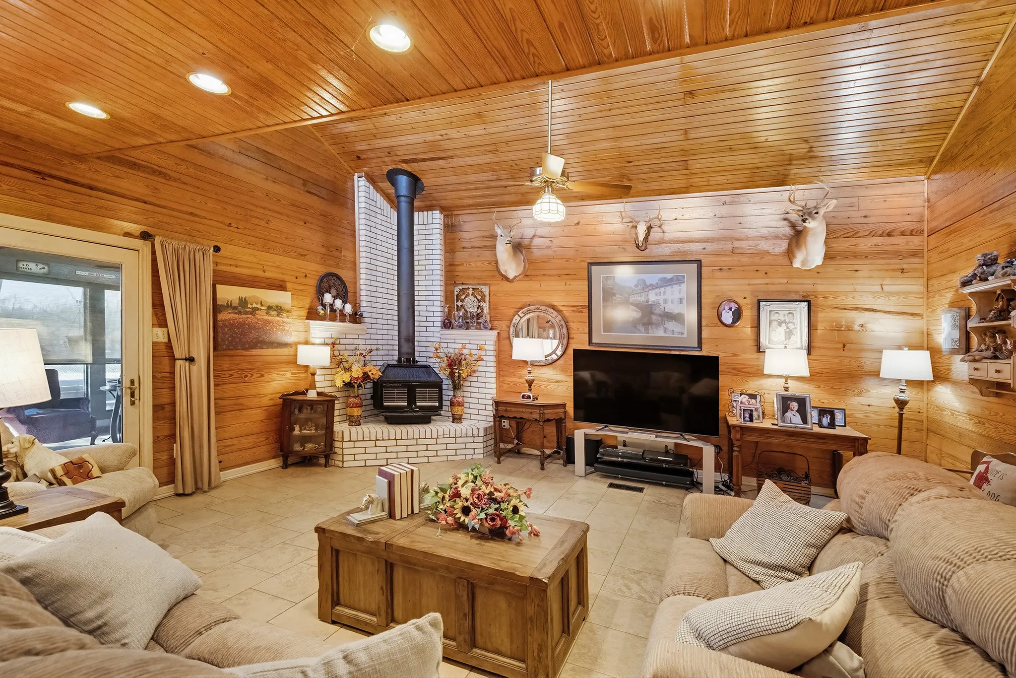 Living room with a wood stove, wood walls, ceiling fan, light tile patterned flooring, and recessed lighting