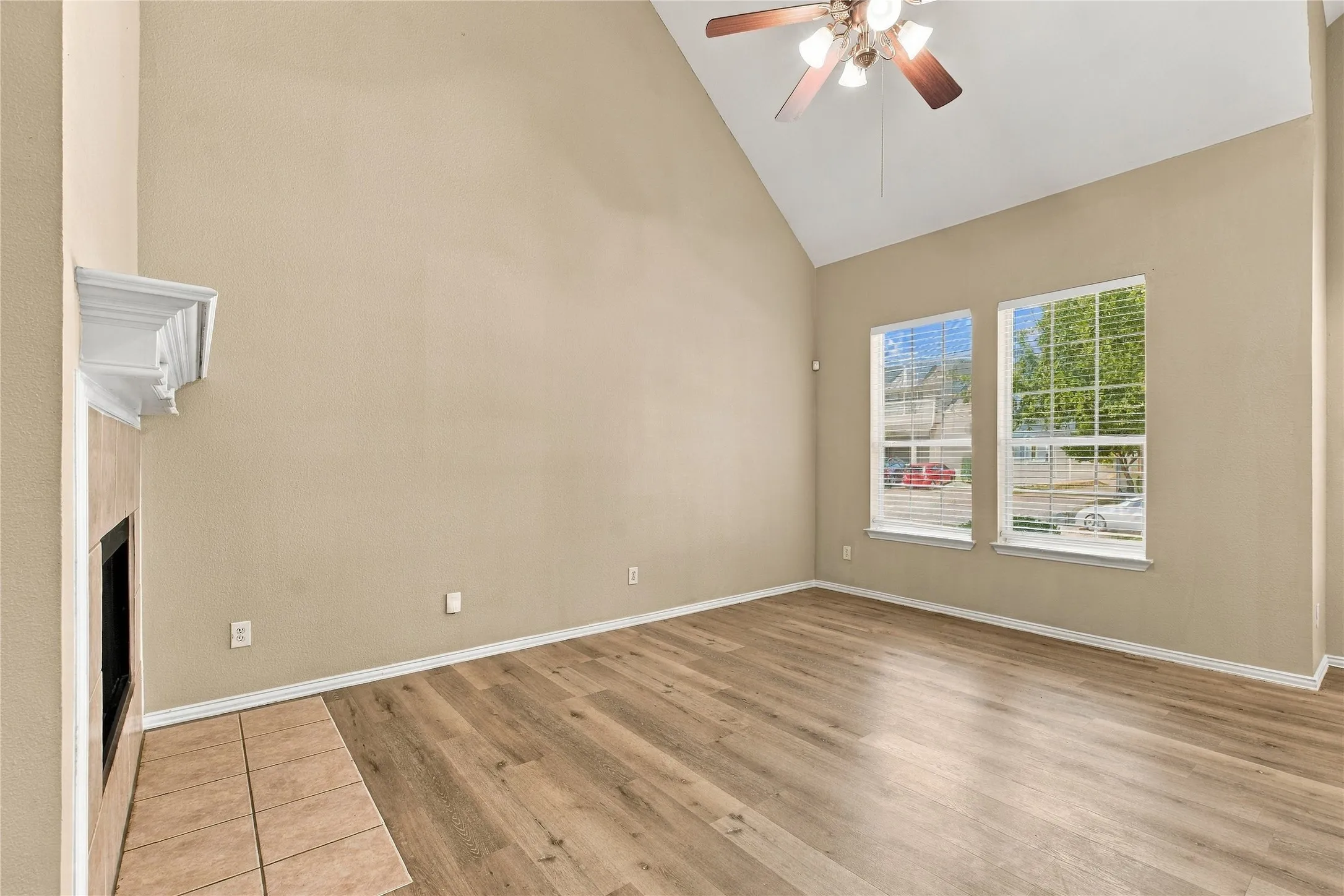 Vaulted ceiling, wood-finish flooring, fireplace with tile surround, two windows with blinds, and a ceiling fan with integrated lighting