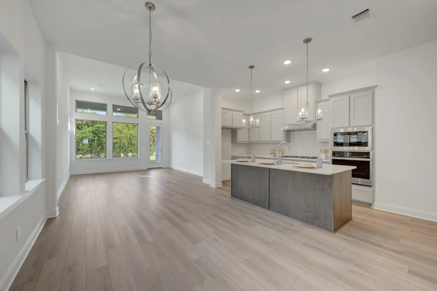 Open-concept living space with light wood-finish flooring, a contemporary chandelier, and large windows