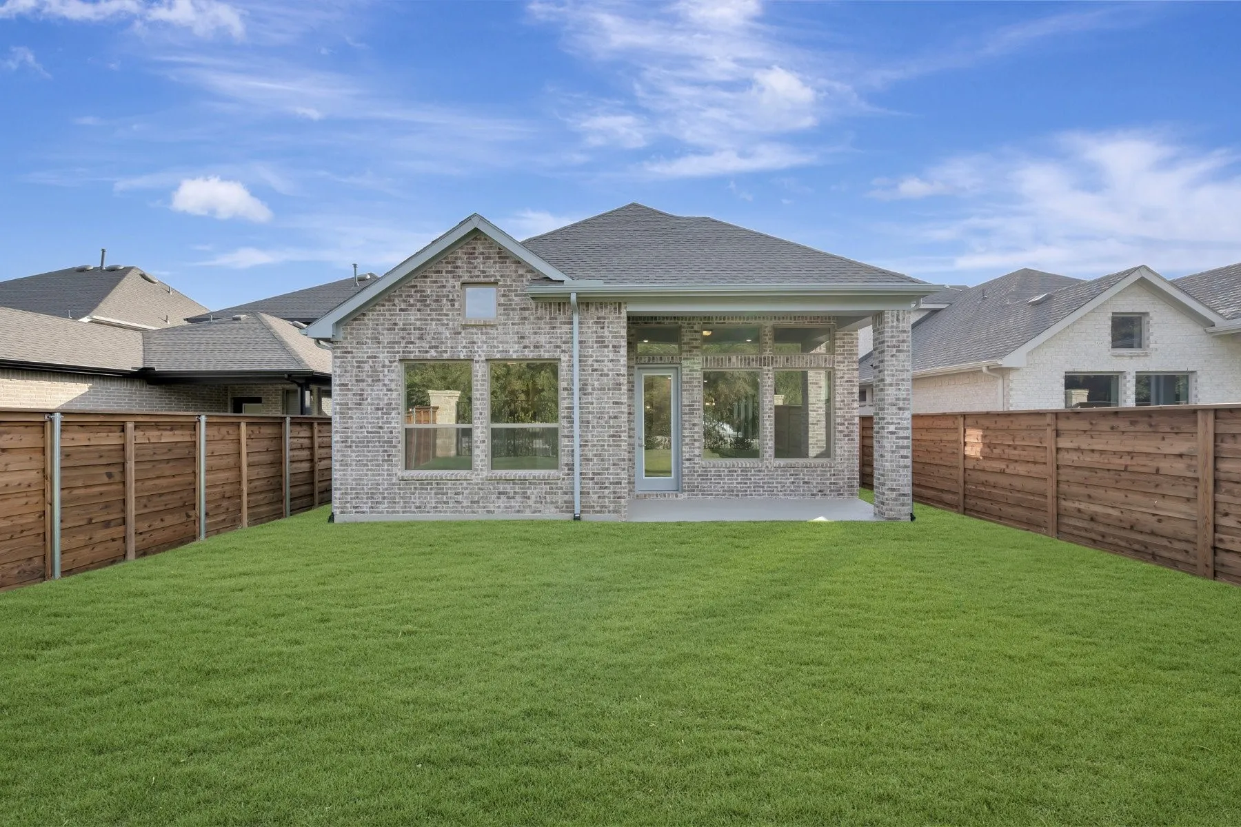 Expansive green lawn, wood fence, covered patio with recessed lighting, large windows, and brick exterior