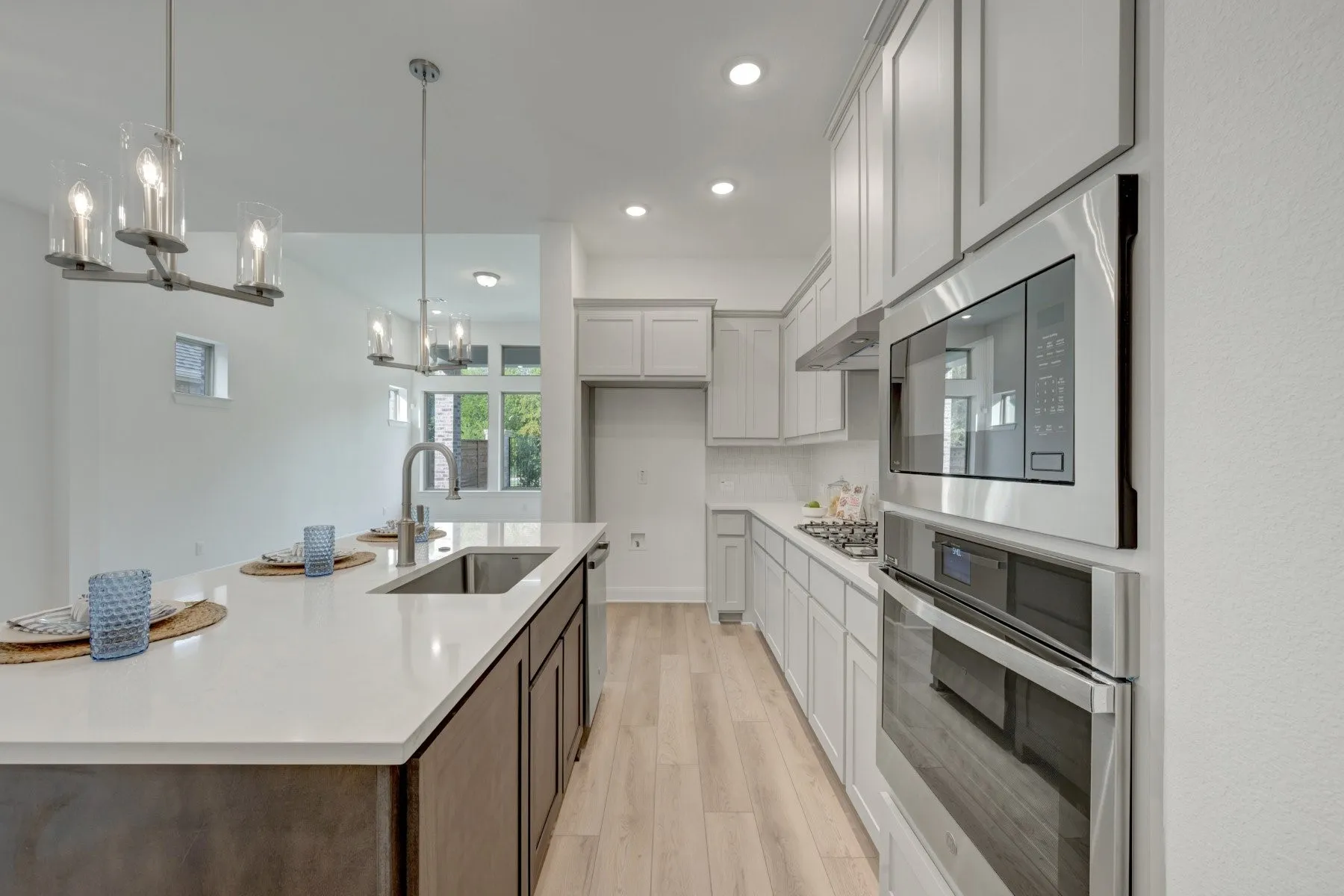 Kitchen featuring an expansive island with a stainless steel sink, wood-finish flooring, light gray cabinetry, and stainless steel appliances including a built-in oven and microwave