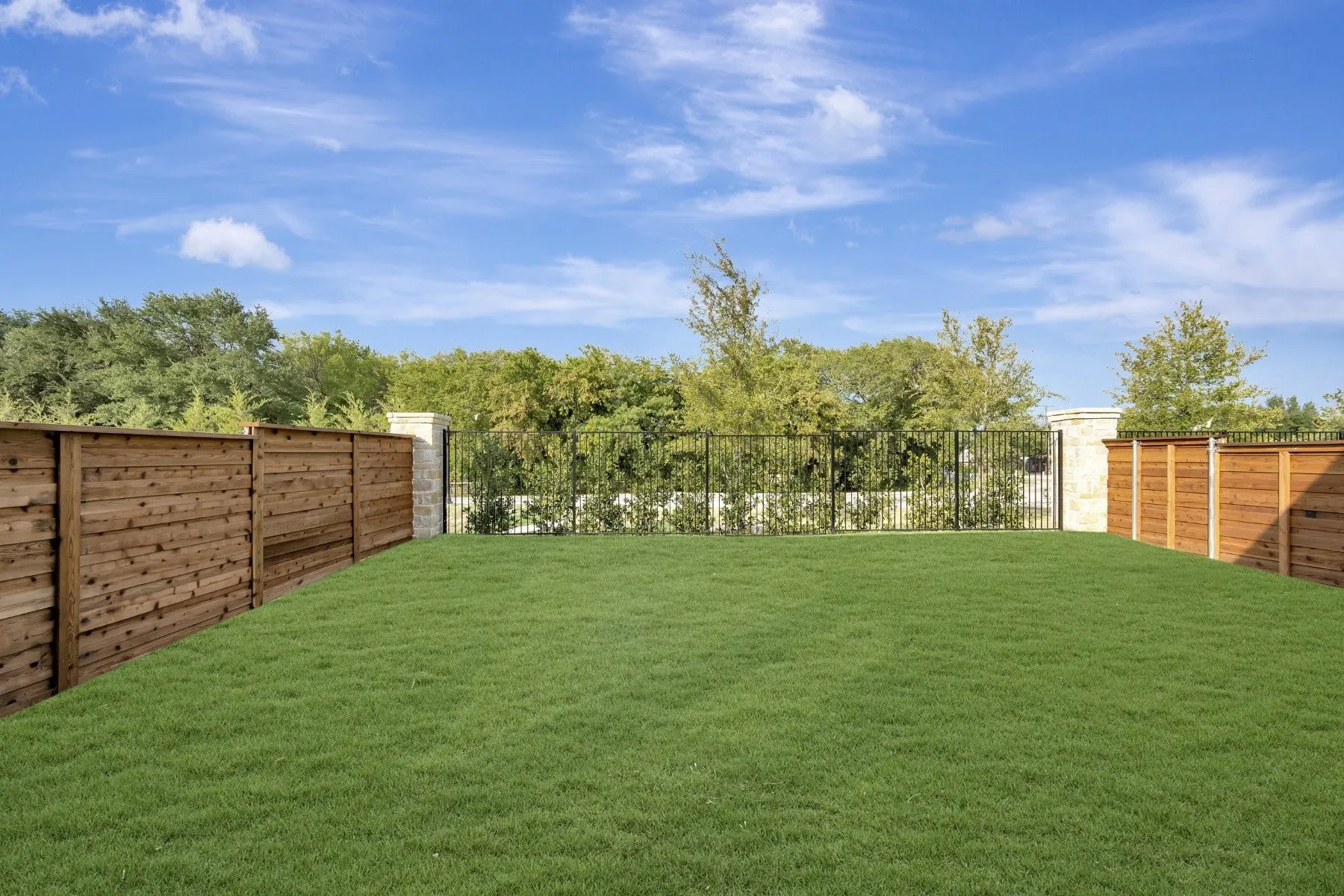 Expansive backyard featuring a manicured lawn, horizontal wood fencing, and black wrought iron fencing with stone pillars