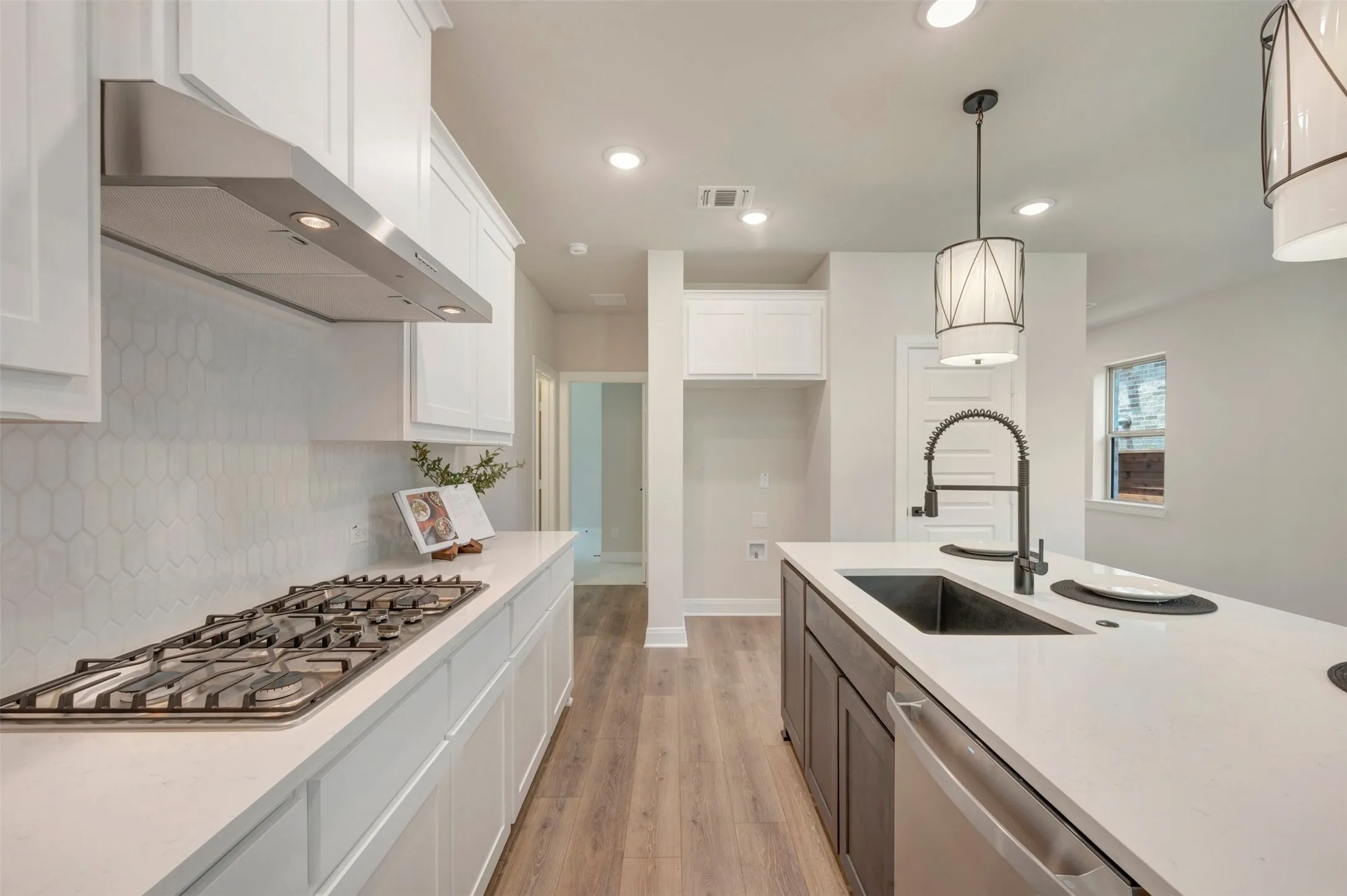 Kitchen featuring white cabinetry, a gas cooktop with stainless steel vent hood, white hexagonal tile backsplash, light wood-finish flooring, and an island with a dark sink and modern pendant lighting