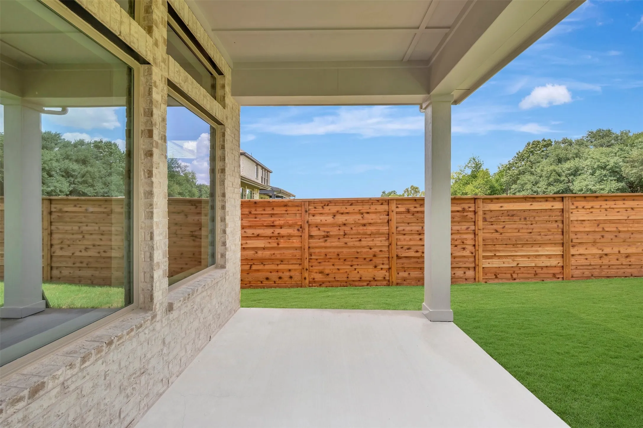 Covered patio with concrete flooring opening to a spacious backyard featuring a manicured lawn, wooden privacy fencing, mature landscaping, and clear blue-sky backdrop