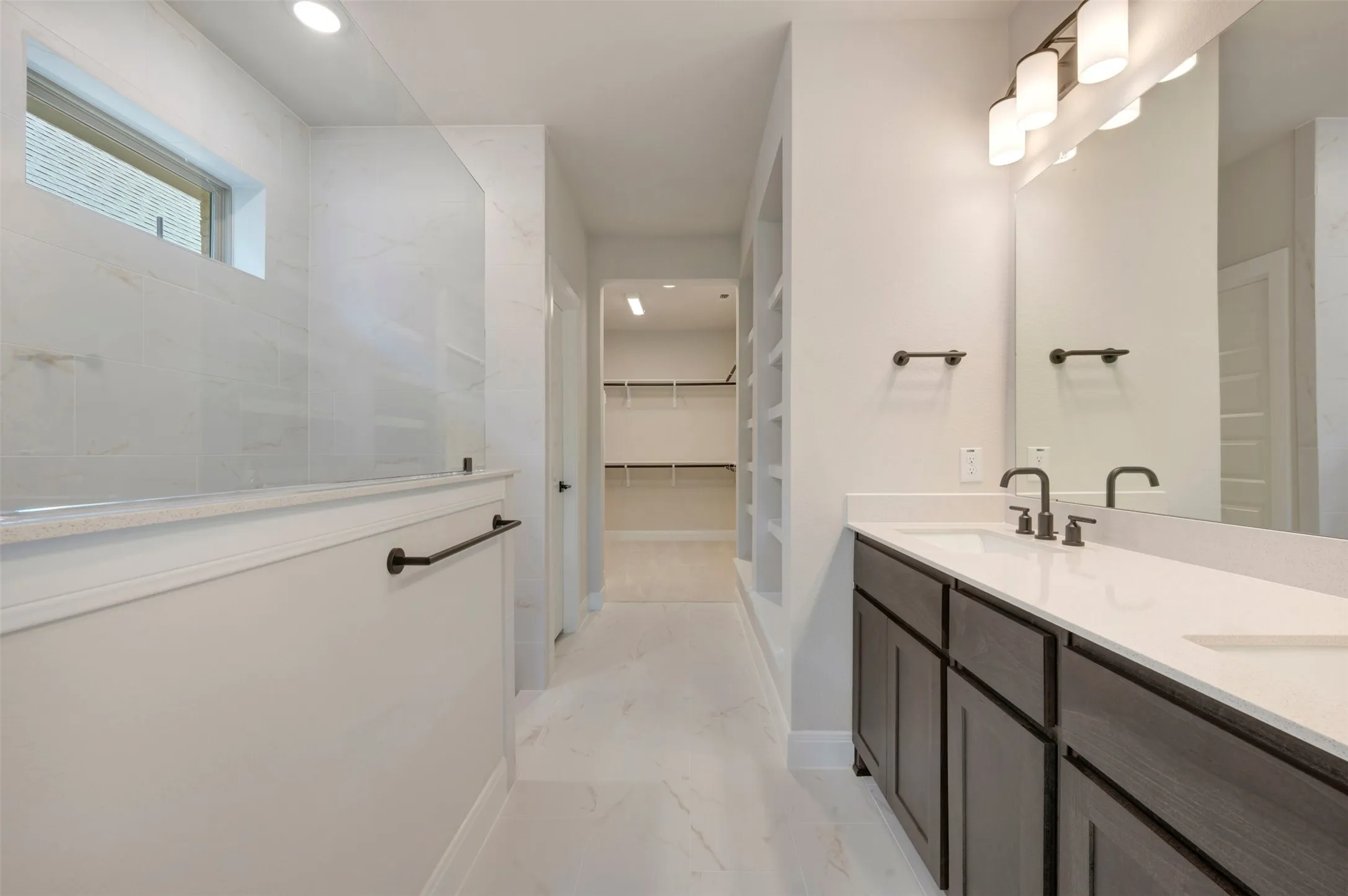 Spa-inspired primary bathroom featuring a spacious walk-in shower with frameless glass enclosure and rainfall showerhead, dual-sink vanity with dark wood cabinetry and brushed nickel fixtures, large framed mirror with integrated lighting, and neutral tile flooring throughout