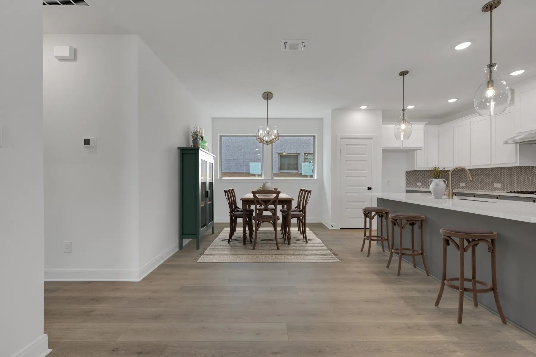 Open-concept living space featuring a modern kitchen with white cabinetry, gray quartz countertops, and stainless steel appliances anchoring one side, while a wood-finish floored dining and living area extends toward expansive windows