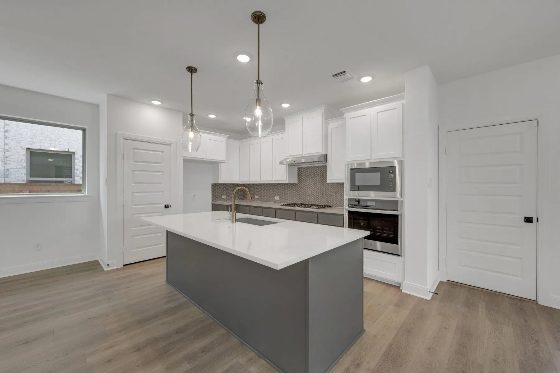 Modern kitchen featuring a large center island with waterfall edge countertop, white cabinetry throughout, stainless steel built-in appliances including wall-mounted oven and microwave, pendant lighting above the island, wood-finish flooring, and ample counter workspace with integrated sink and prep areas