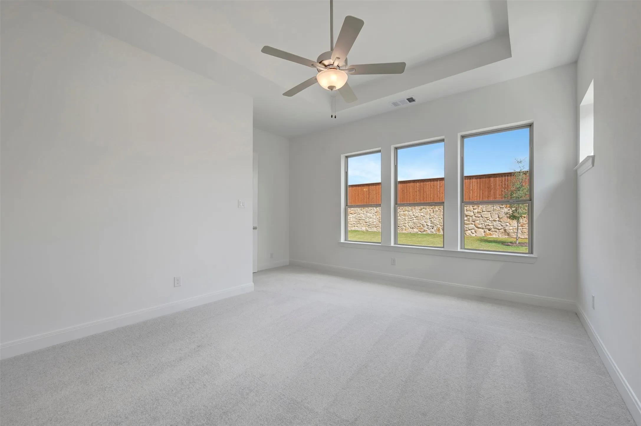 Tray ceiling with a ceiling fan, three large windows, light-colored carpeting, and a high window