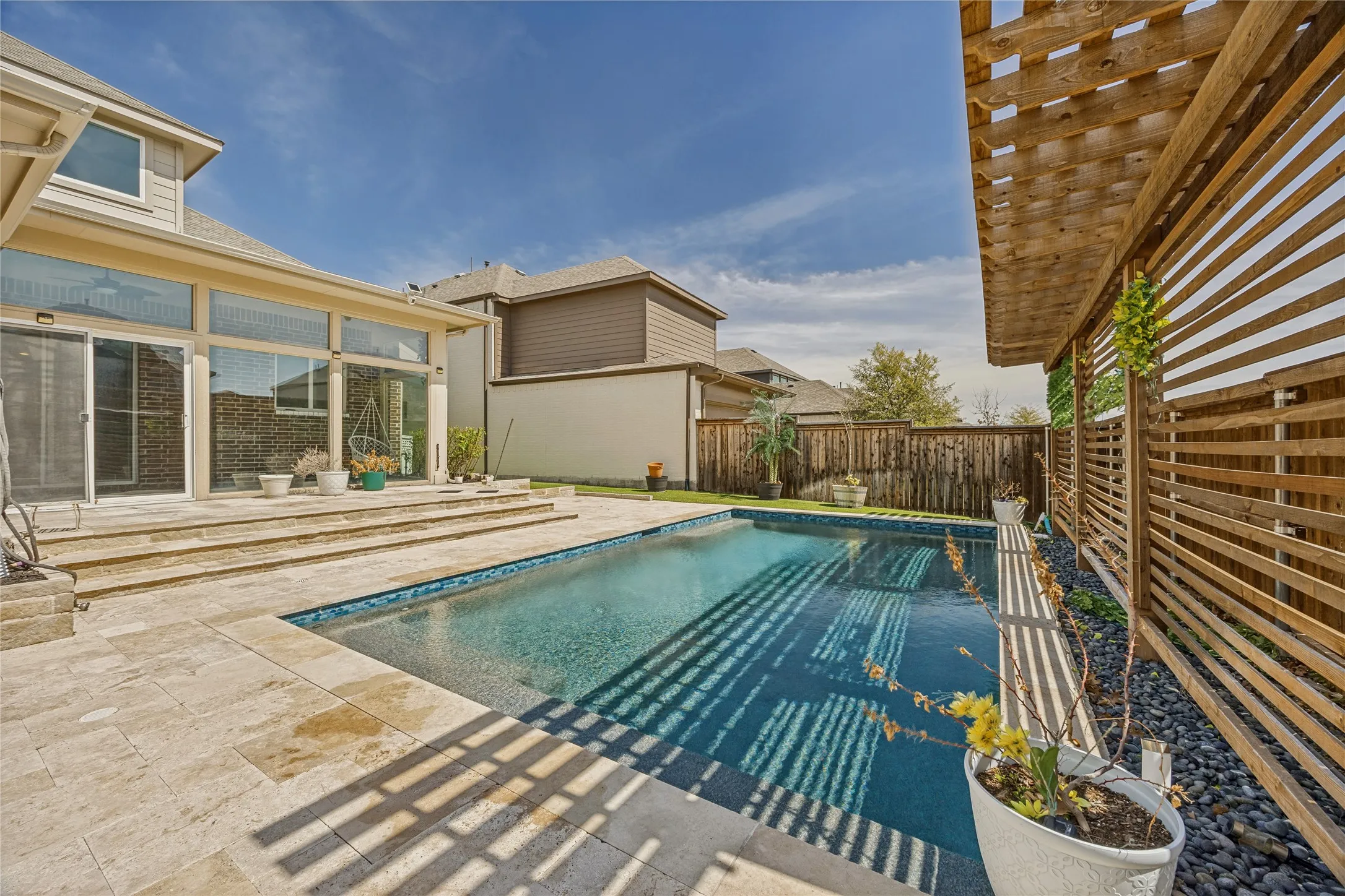 Private backyard oasis featuring a rectangular swimming pool with tiled coping, a multi-tiered patio with stone-finish pavers, and full-height glass doors along the rear facade