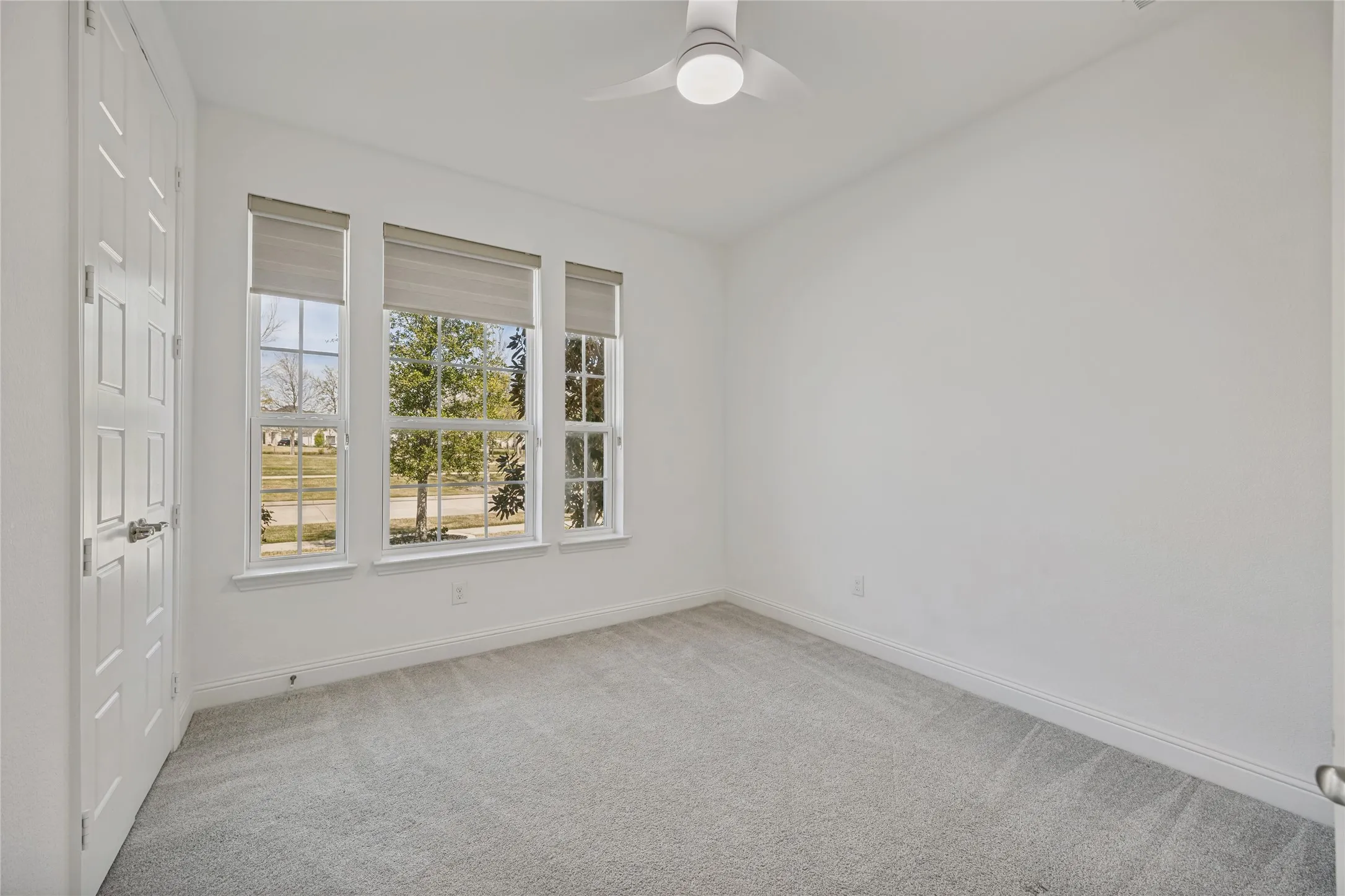 Light-filled interior space featuring three large windows with roller shades, neutral carpet flooring, and a modern ceiling fan