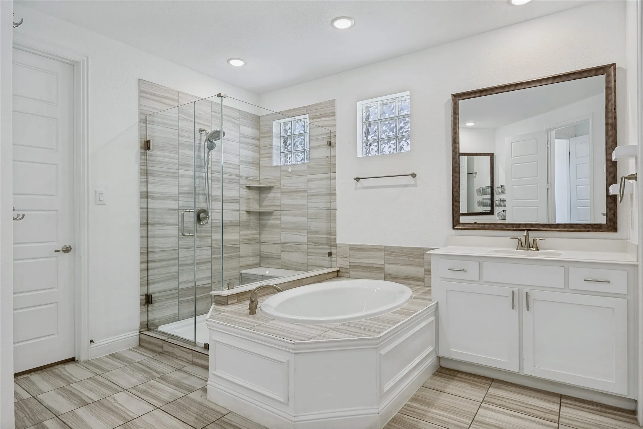 Tiled flooring throughout this bathroom featuring a built-in soaking tub, a glass-enclosed shower with a tiled surround, a single vanity with a white countertop, and a large framed mirror
