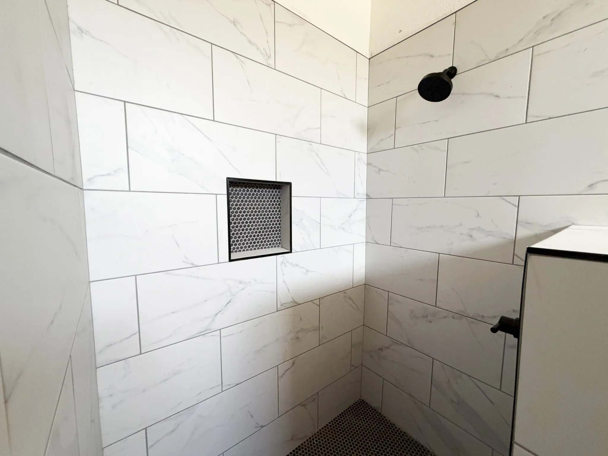 Shower with white rectangular wall tiles featuring subtle veining, black circular mosaic floor tiles, and a recessed niche with a black grate