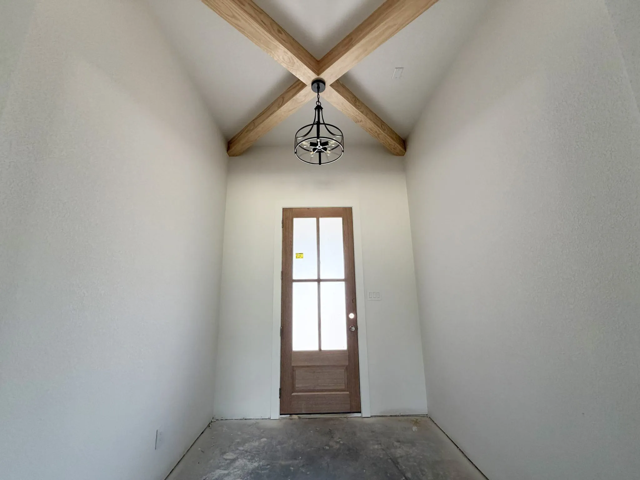 Vaulted ceiling with exposed wood beams, modern black chandelier, wood-finish door with glass panels, and a concrete-finish floor