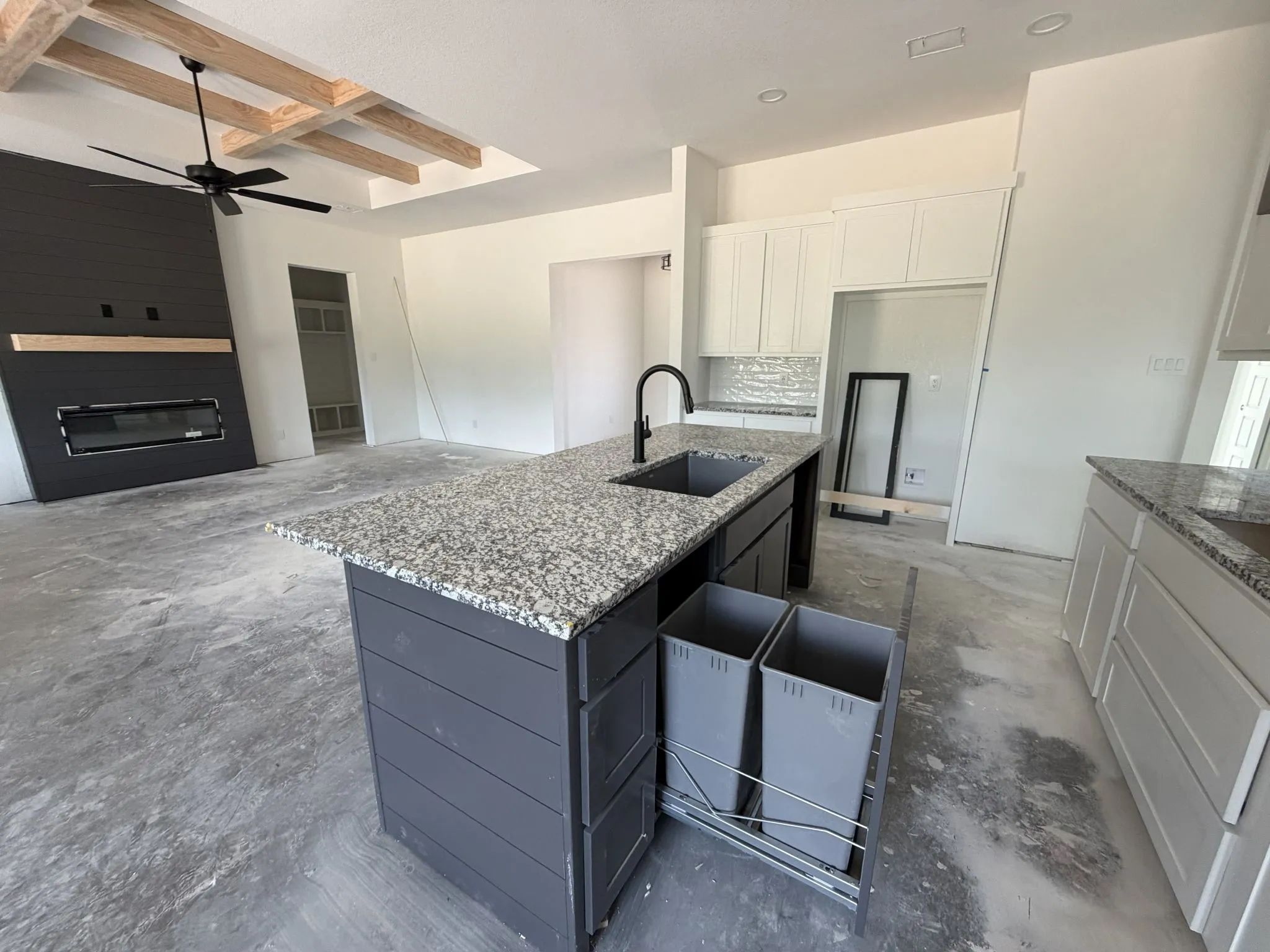 Granite countertop island with integrated sink, pull-out waste bins, white cabinetry, and shiplap fireplace with wood mantel