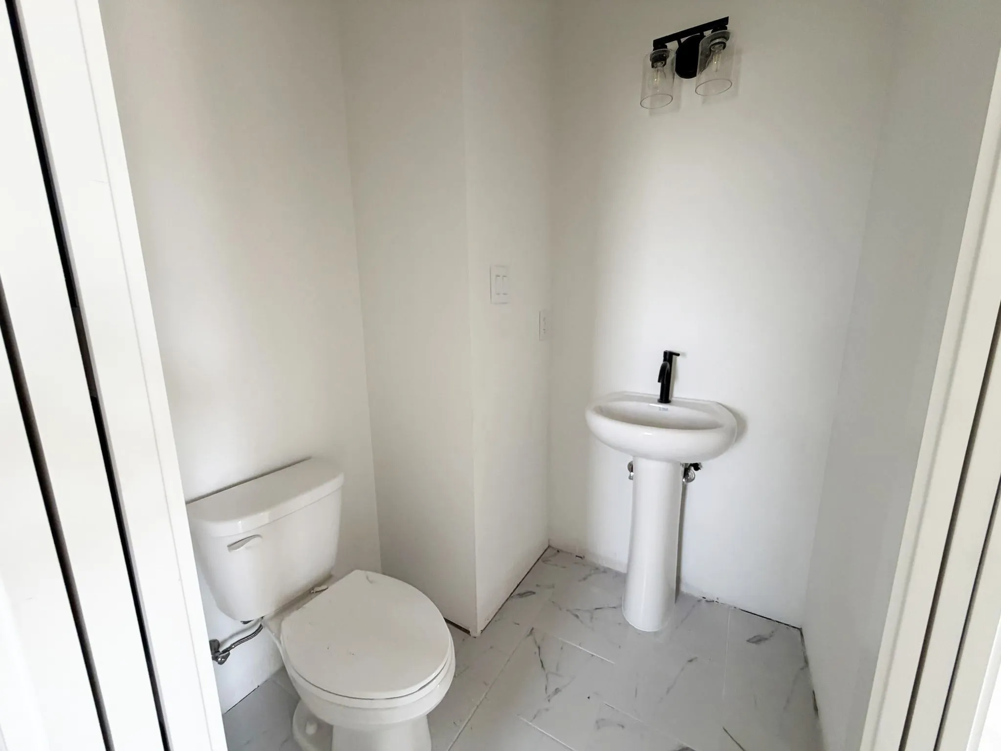 White pedestal sink with a matte black faucet, ceramic floor tiling, and a two-bulb wall-mounted light fixture