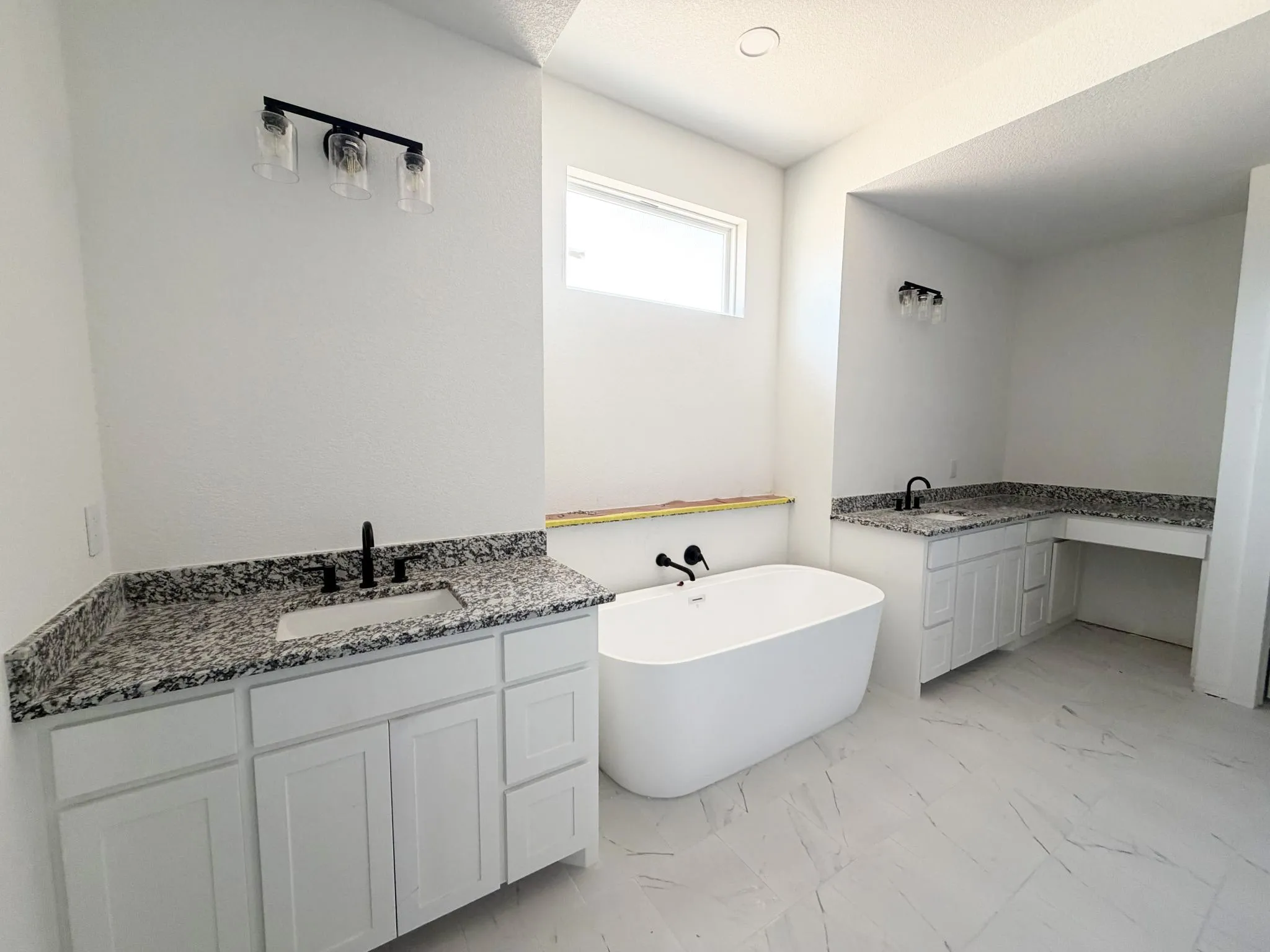 Freestanding soaking tub, double vanity with granite countertops, white cabinetry, matte black fixtures, and large-format tile flooring