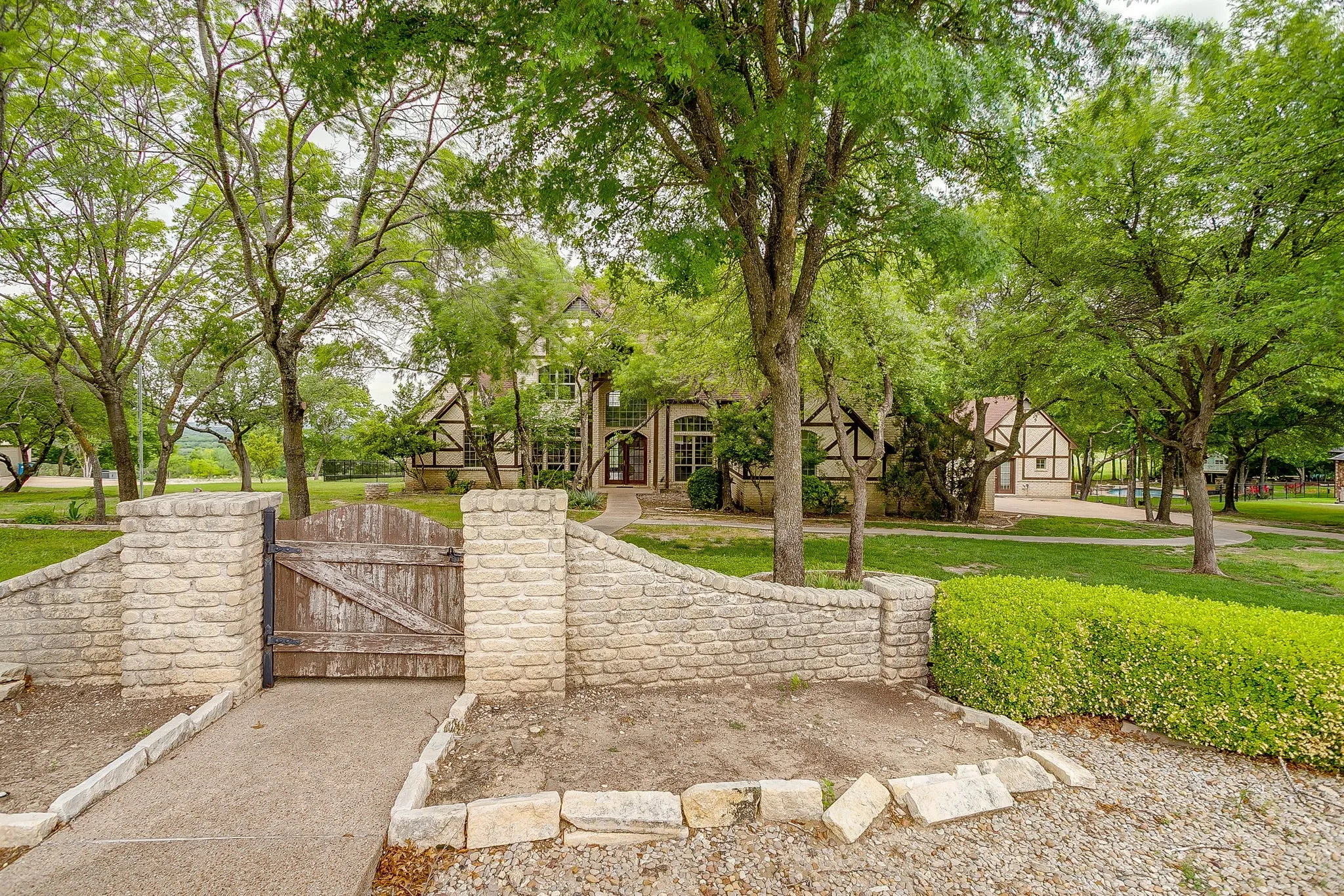 The property features a stone wall entrance with a wooden gate, a paved pathway, and mature trees providing natural shade