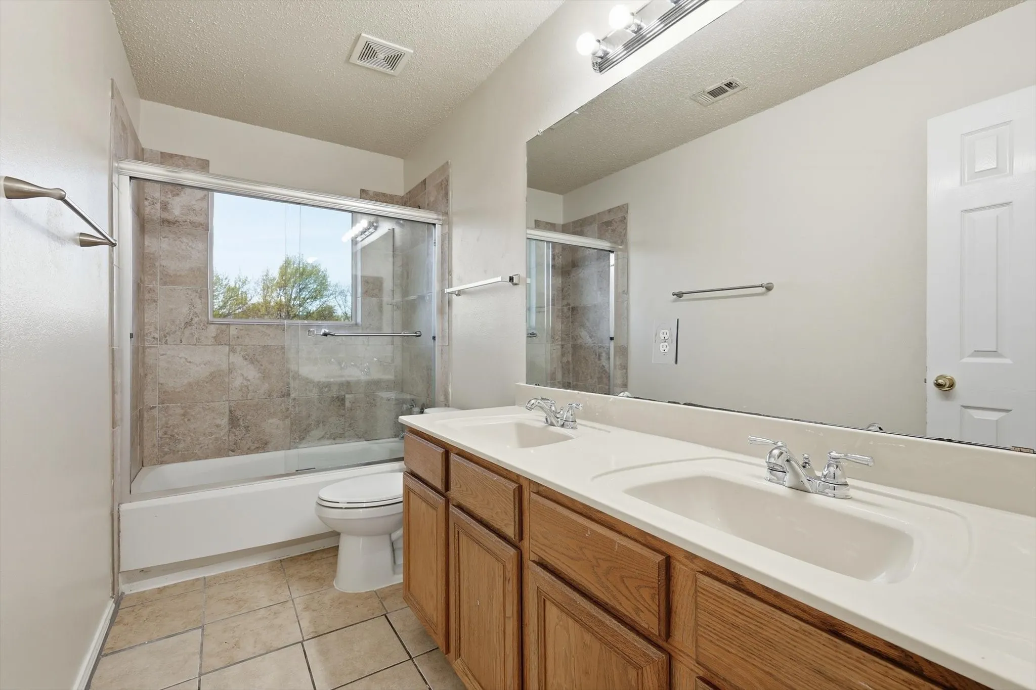 Bathroom with double vanity, a textured ceiling, shower / bath combination with glass door, and light tile patterned floors