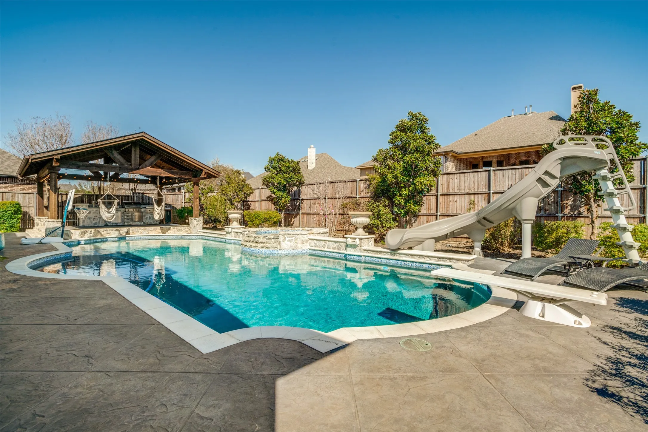 Resort oasis pool with in-pool barstools, a spa, a water slide, a diving board, and a tanning bed next to the outdoor kitchen and pergola