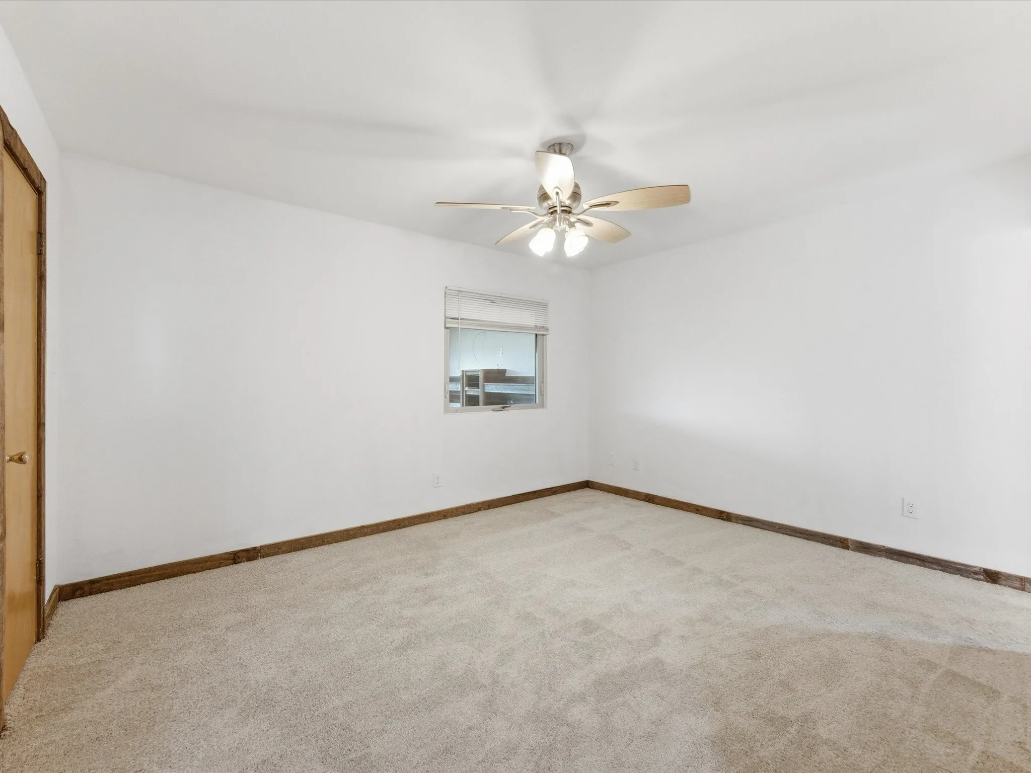 Spacious room featuring neutral carpeting, white walls, a single window with blinds, a ceiling fan with integrated lighting, and wood-finish trim