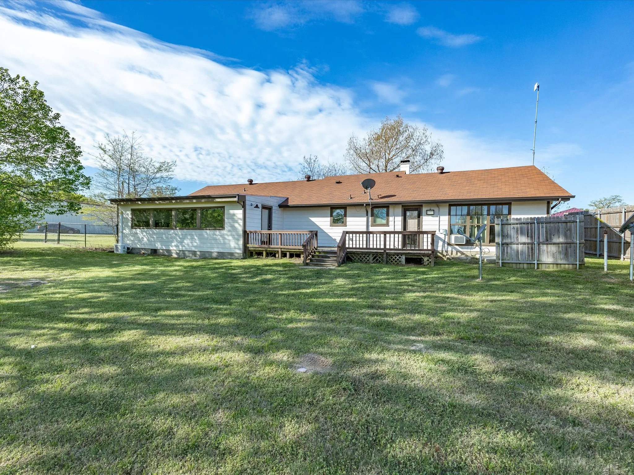 Expansive backyard featuring a manicured lawn, a chain-link fence, a brown shingle roof, a white exterior, and a wood deck
