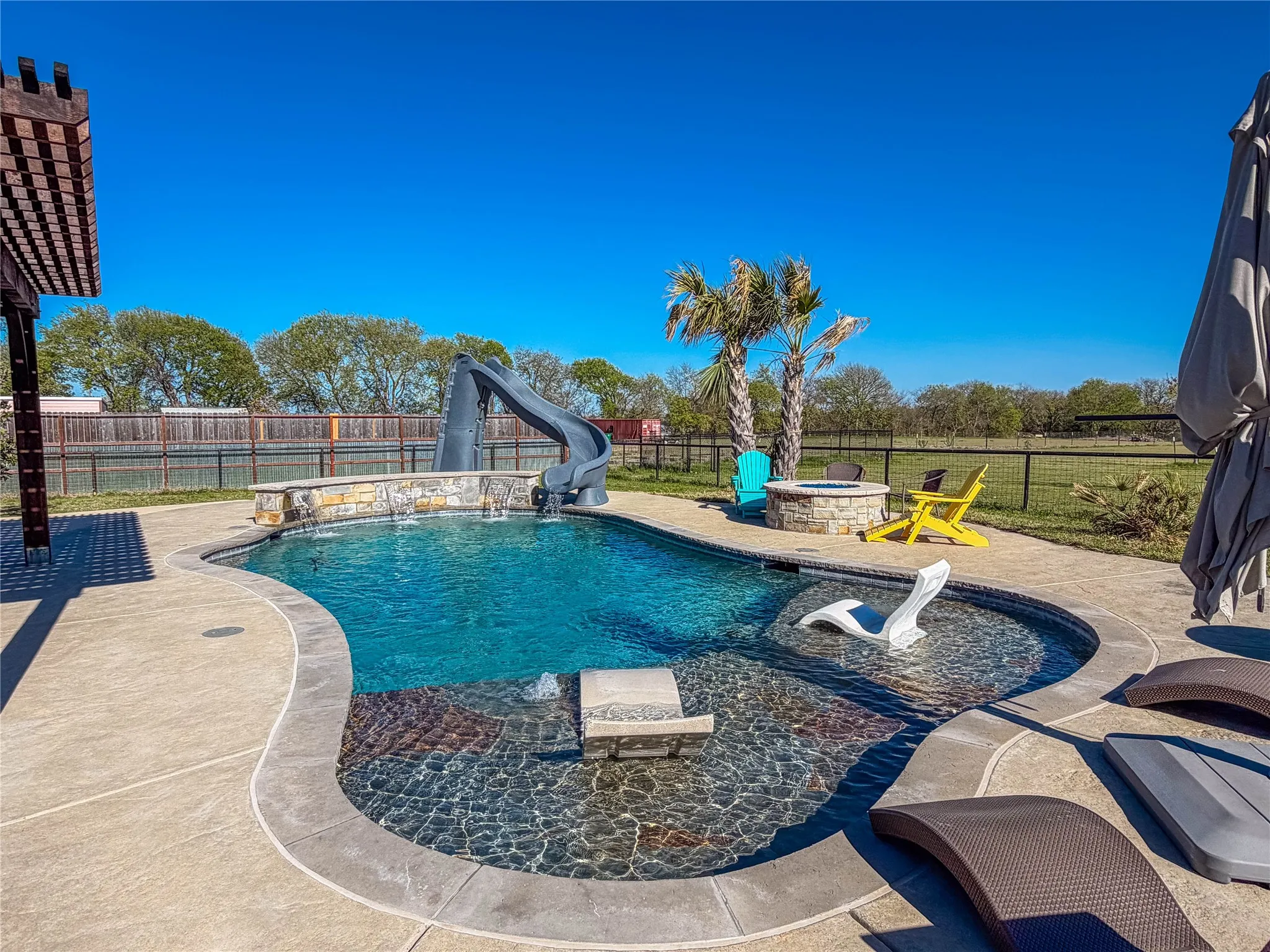 View of pool featuring a water slide, patio surround, a fenced backyard, and a fire pit