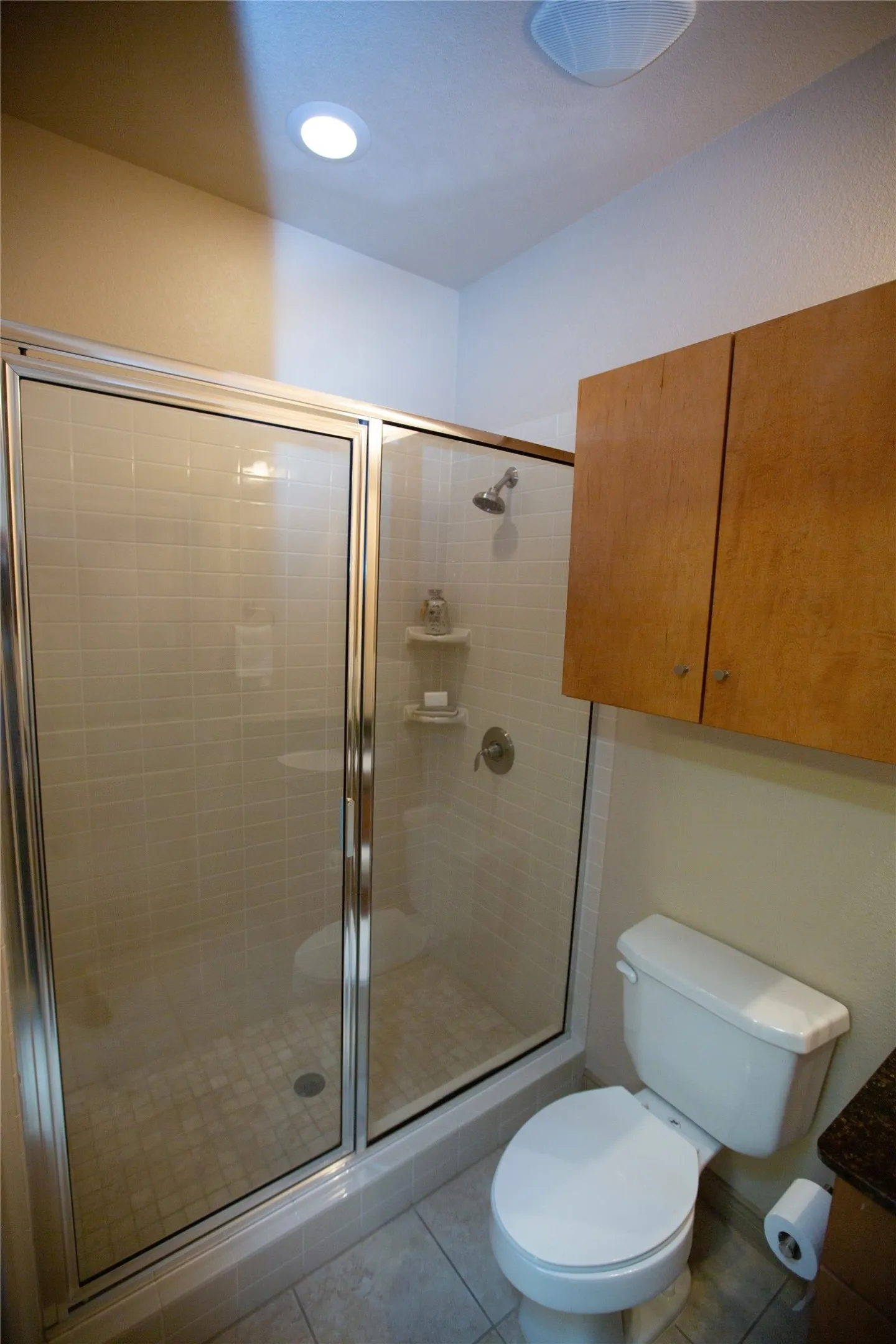 Shower enclosure with glass doors and chrome frame, subway tile surround, tiled floor, white commode, and wood-finish cabinetry