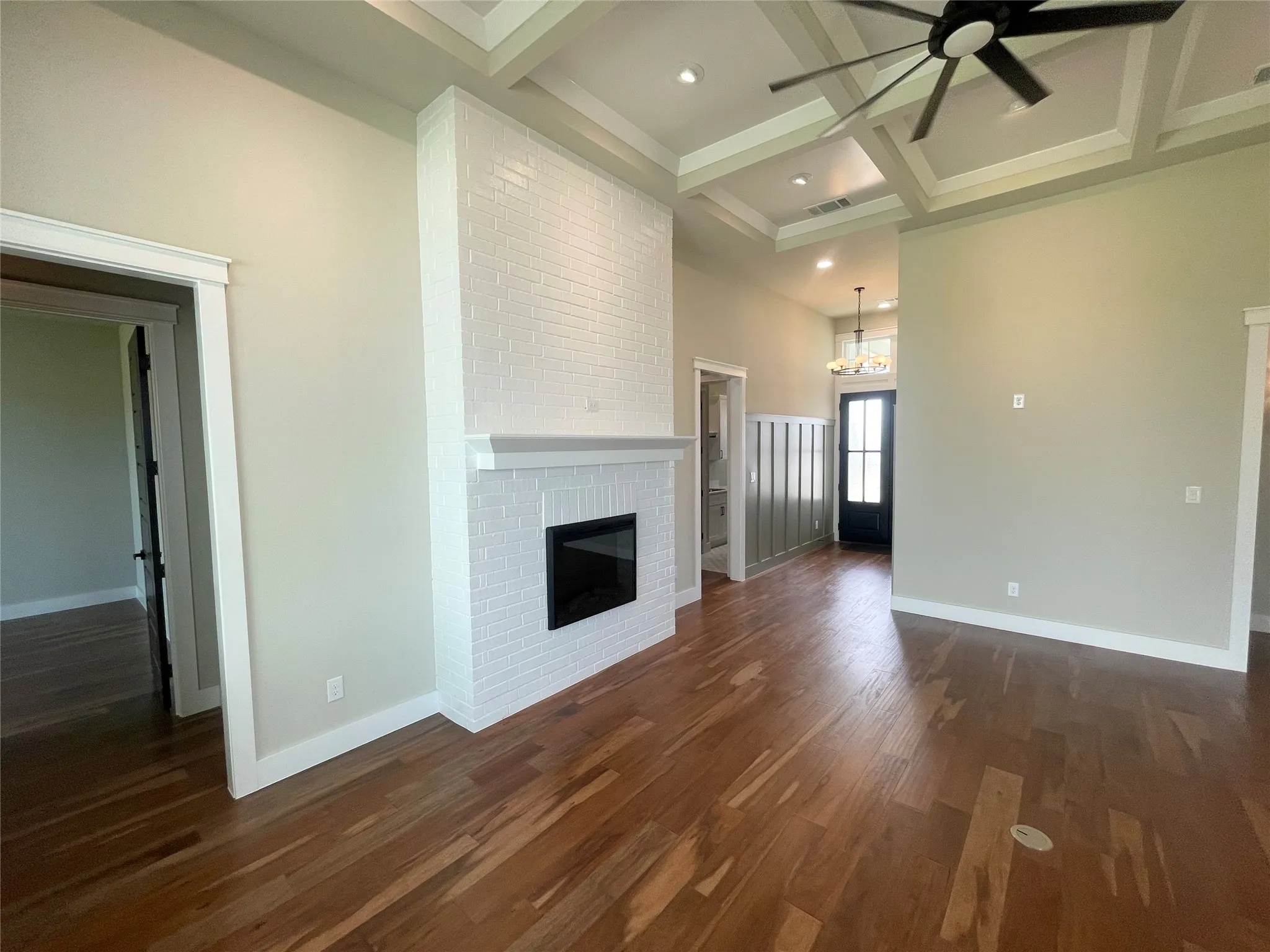 Hardwood flooring throughout, a white brick fireplace, a coffered ceiling with a ceiling fan, and a paneled entry wall