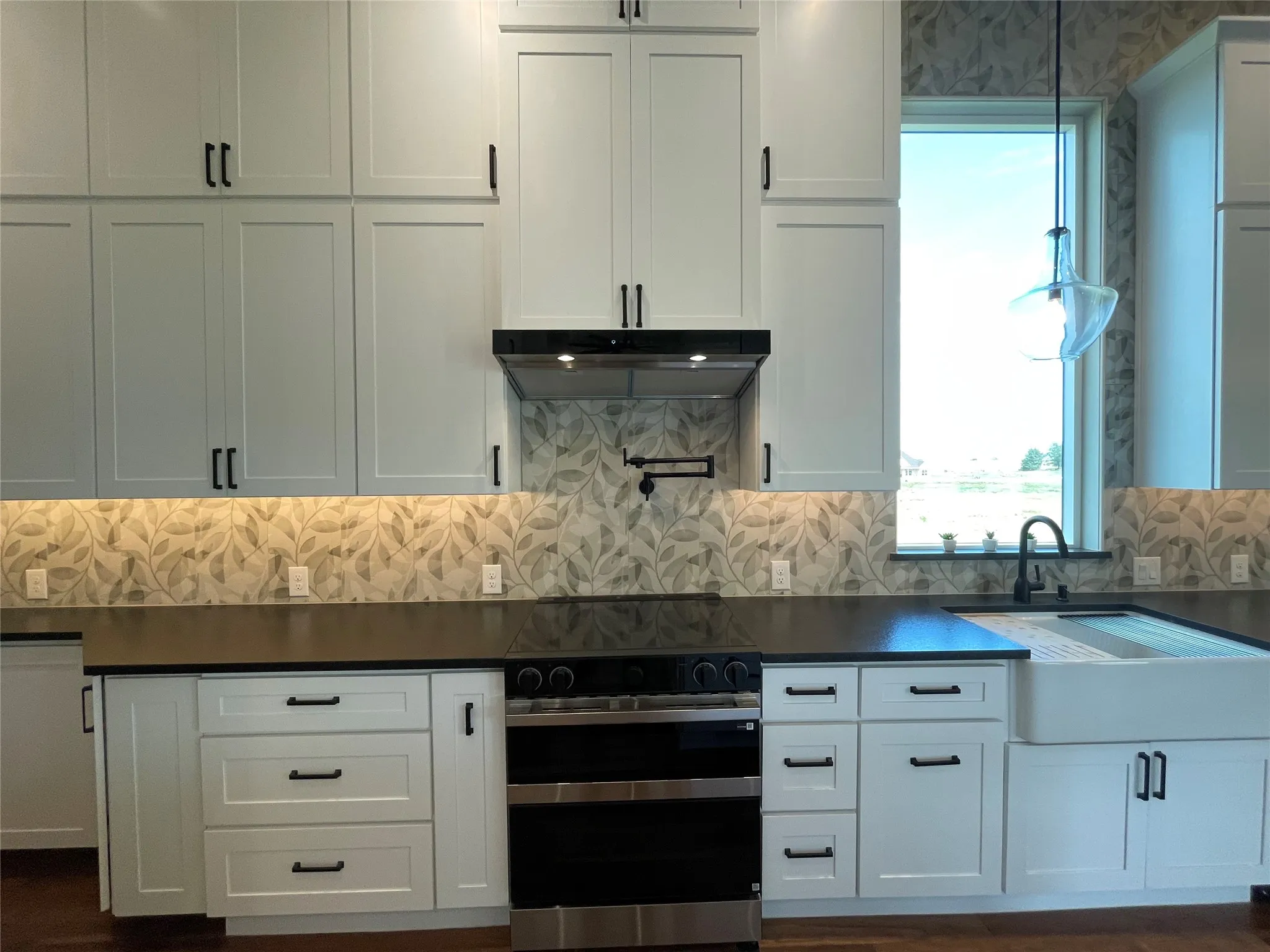 White shaker-style cabinetry with black hardware, patterned tile backsplash, dark countertops, and a farmhouse sink