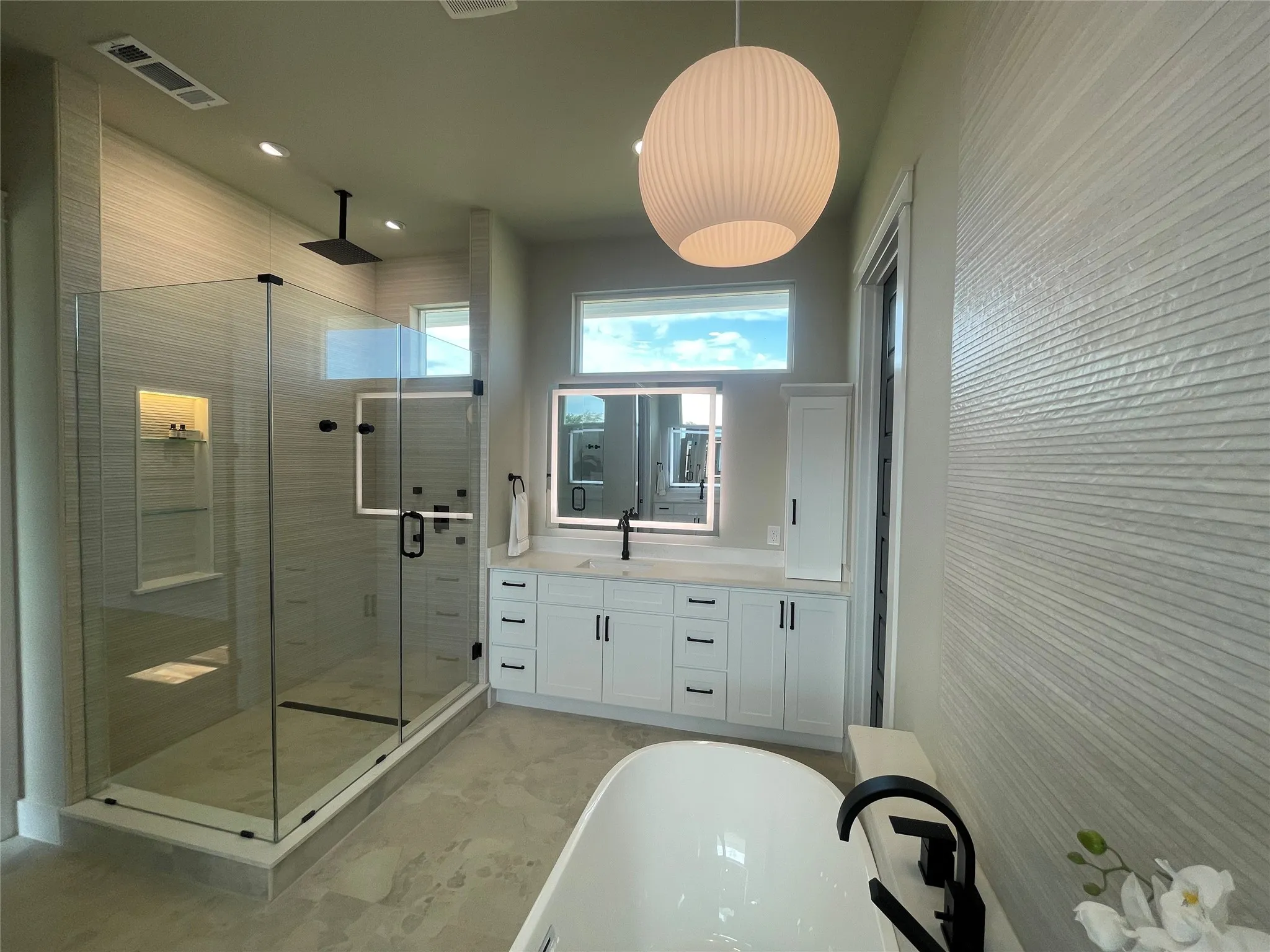 Glass-enclosed shower with a rainfall showerhead, freestanding soaking tub, dual-sink vanity with white cabinetry, and an illuminated mirror