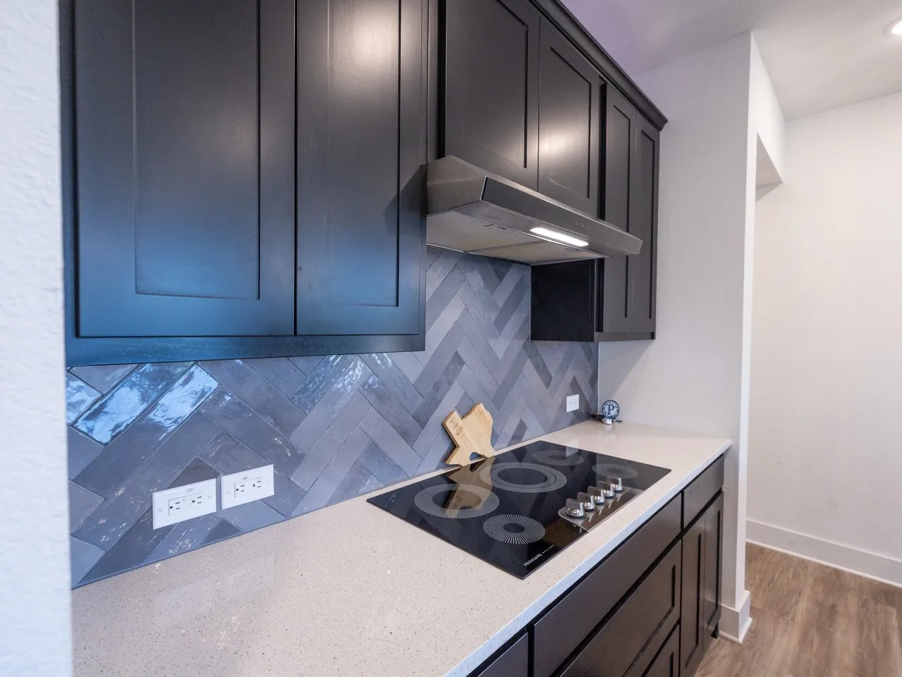 Black shaker-style cabinetry, herringbone tile backsplash, integrated cooktop, light-toned countertop, and wood-finish flooring