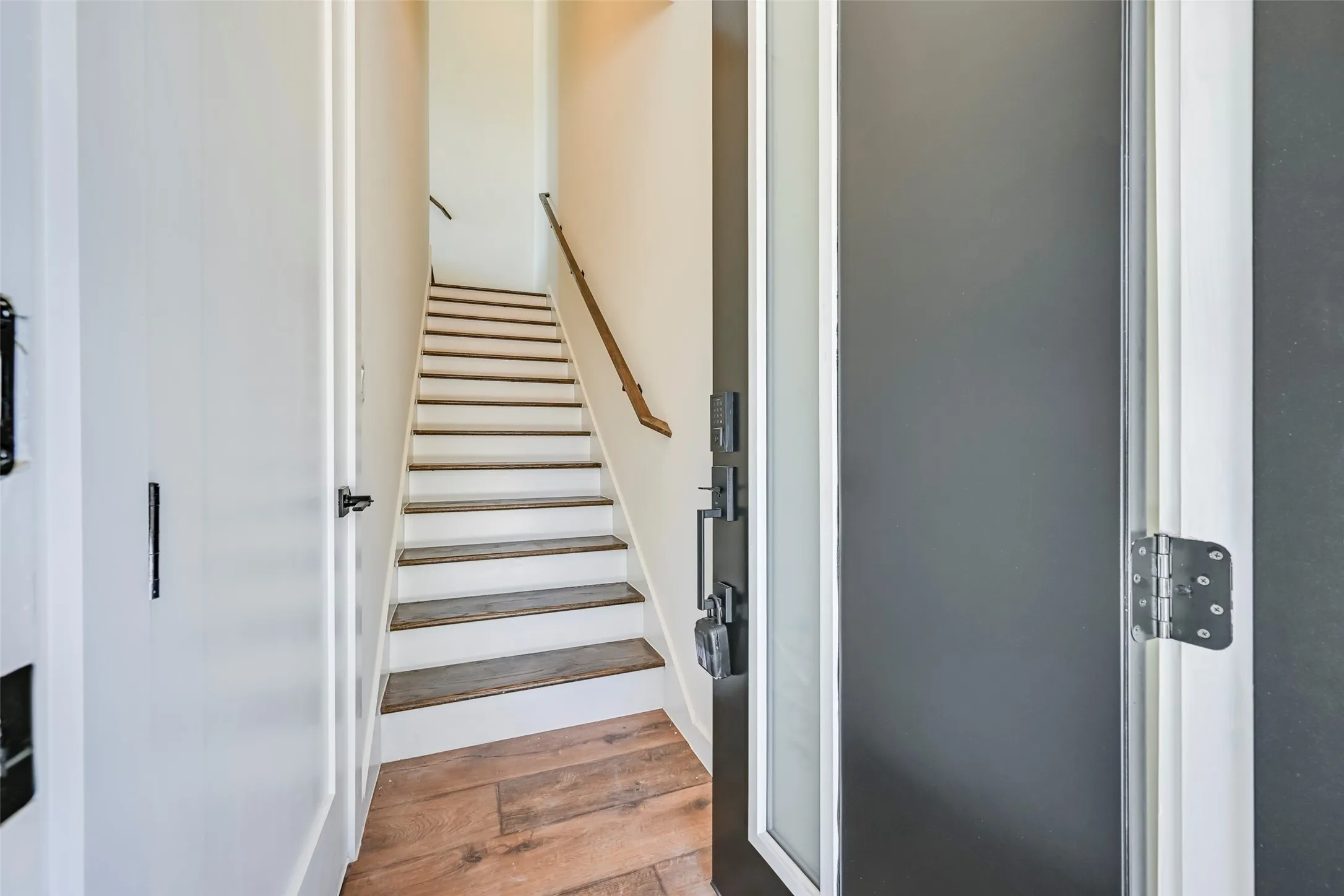 Wood-finish flooring, white paneled door, black hardware, wood handrail, and white stair risers