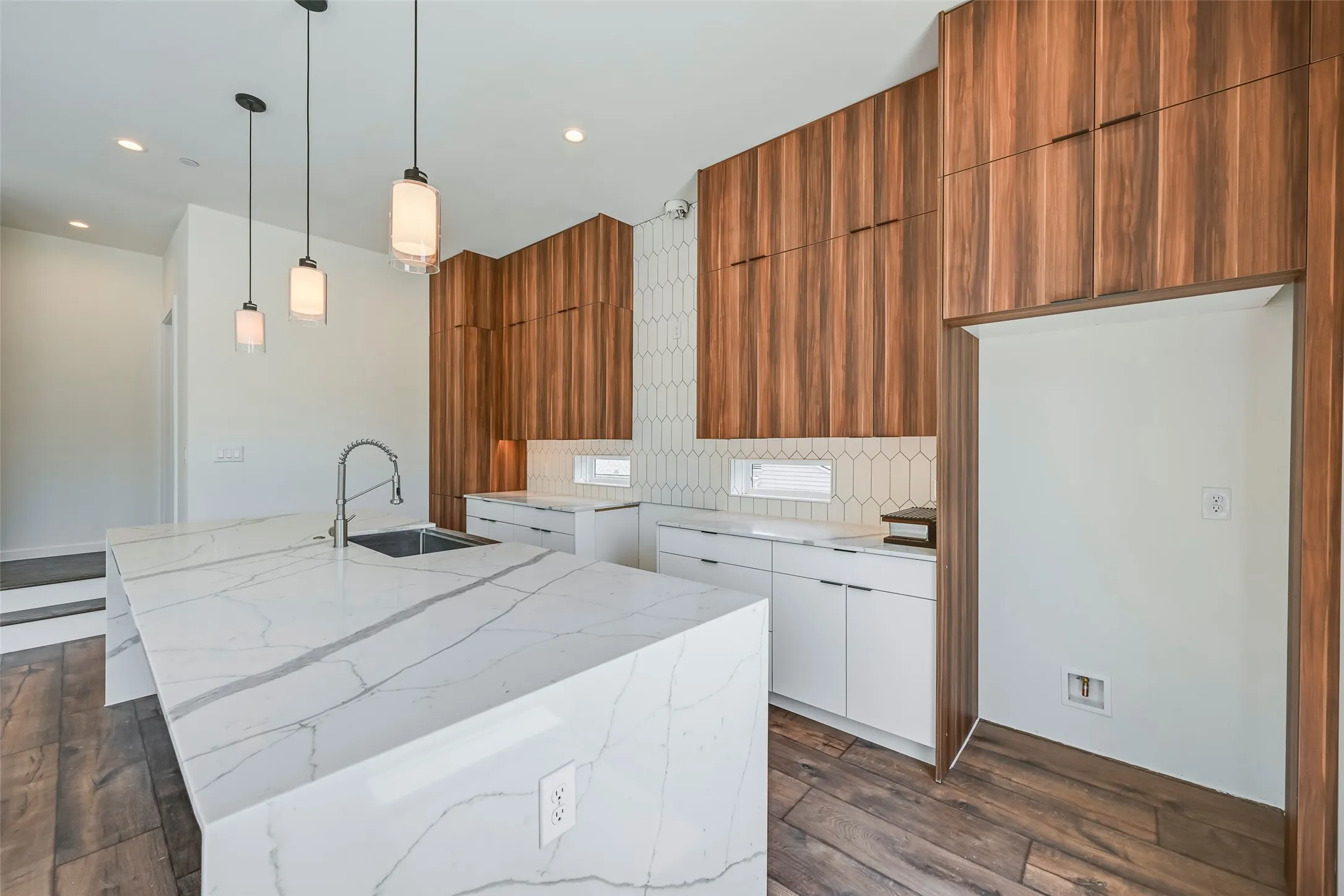 Waterfall island with integrated sink and chrome faucet, custom wood-finish cabinetry, white flat-panel lower cabinetry, geometric tile backsplash, and recessed and pendant lighting