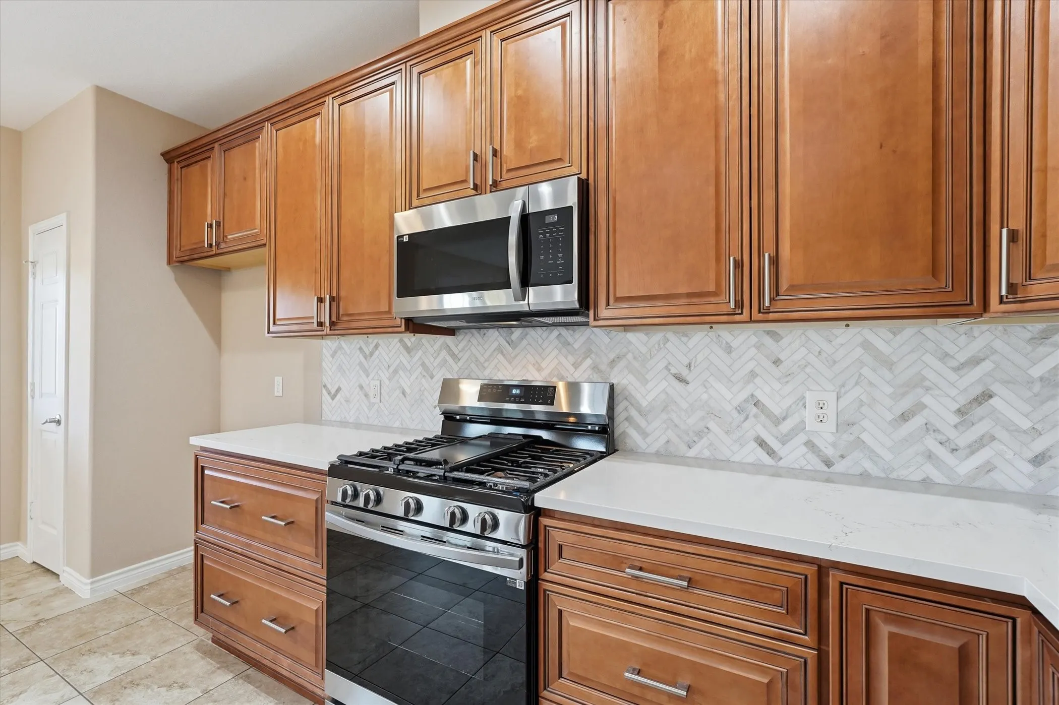 Kitchen with stainless steel appliances, wood finish cabinets, backsplash, light tile patterned floors, and light stone counters
