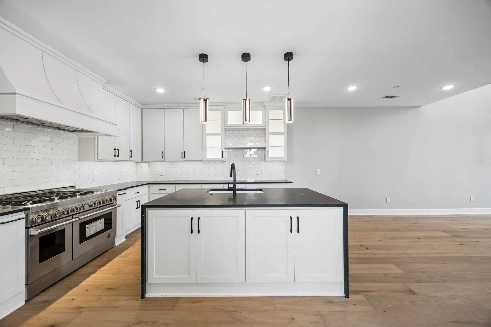 Kitchen with a center island with sink, range with two ovens, light wood finished floors, white cabinetry, and hanging light fixtures