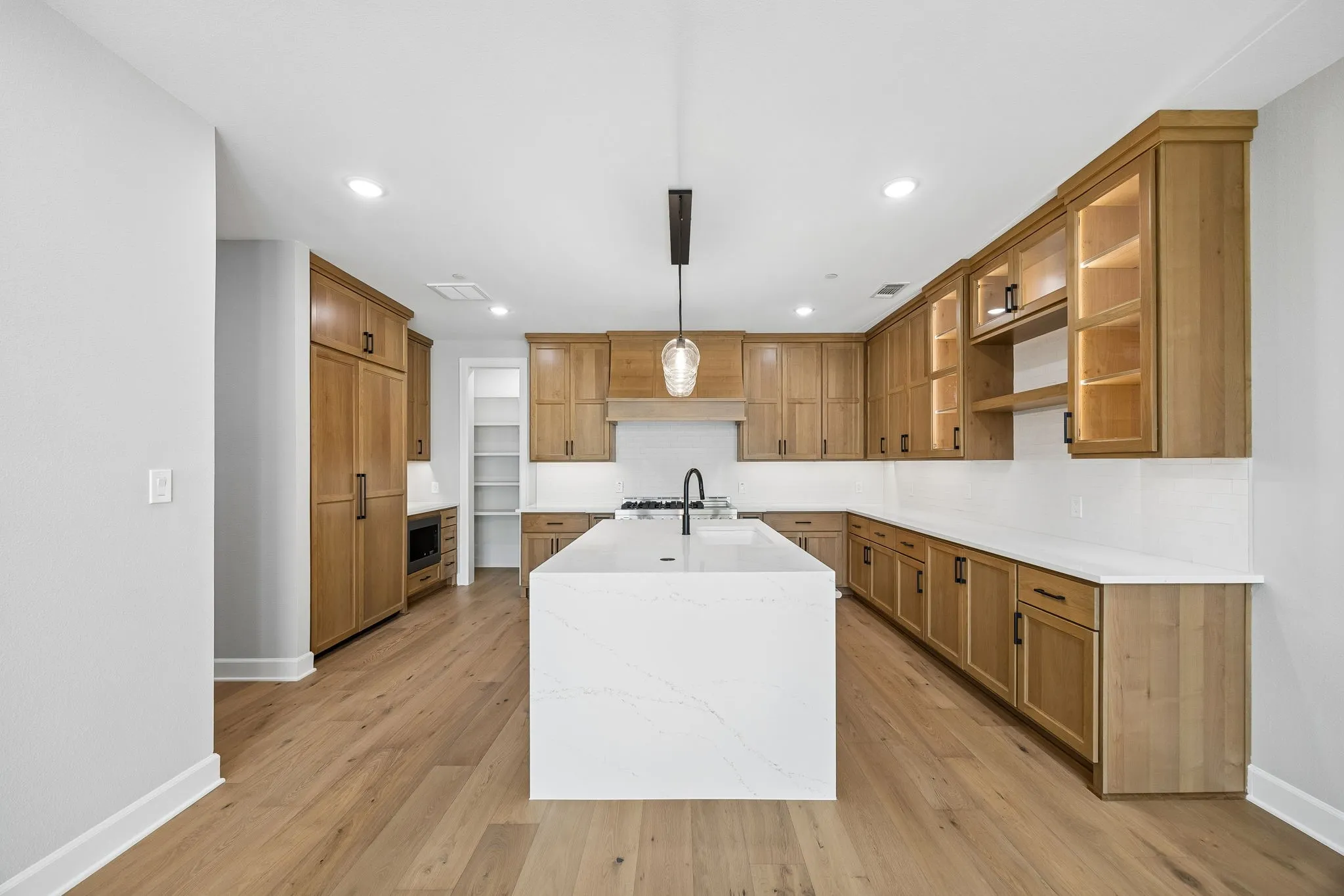 Kitchen with a center island with sink, light stone counters, hanging light fixtures, wood finish cabinetry, and decorative backsplash