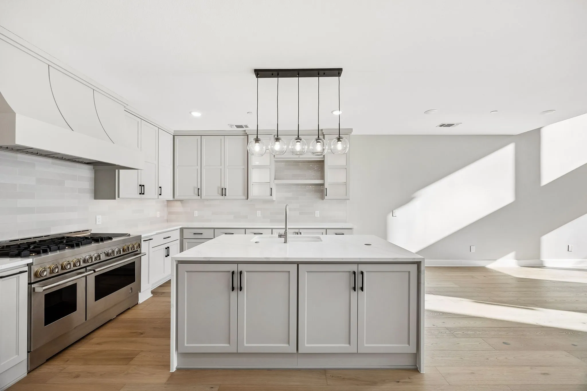 Kitchen with a center island with sink, range with two ovens, open shelves, light wood-type flooring, and light stone countertops