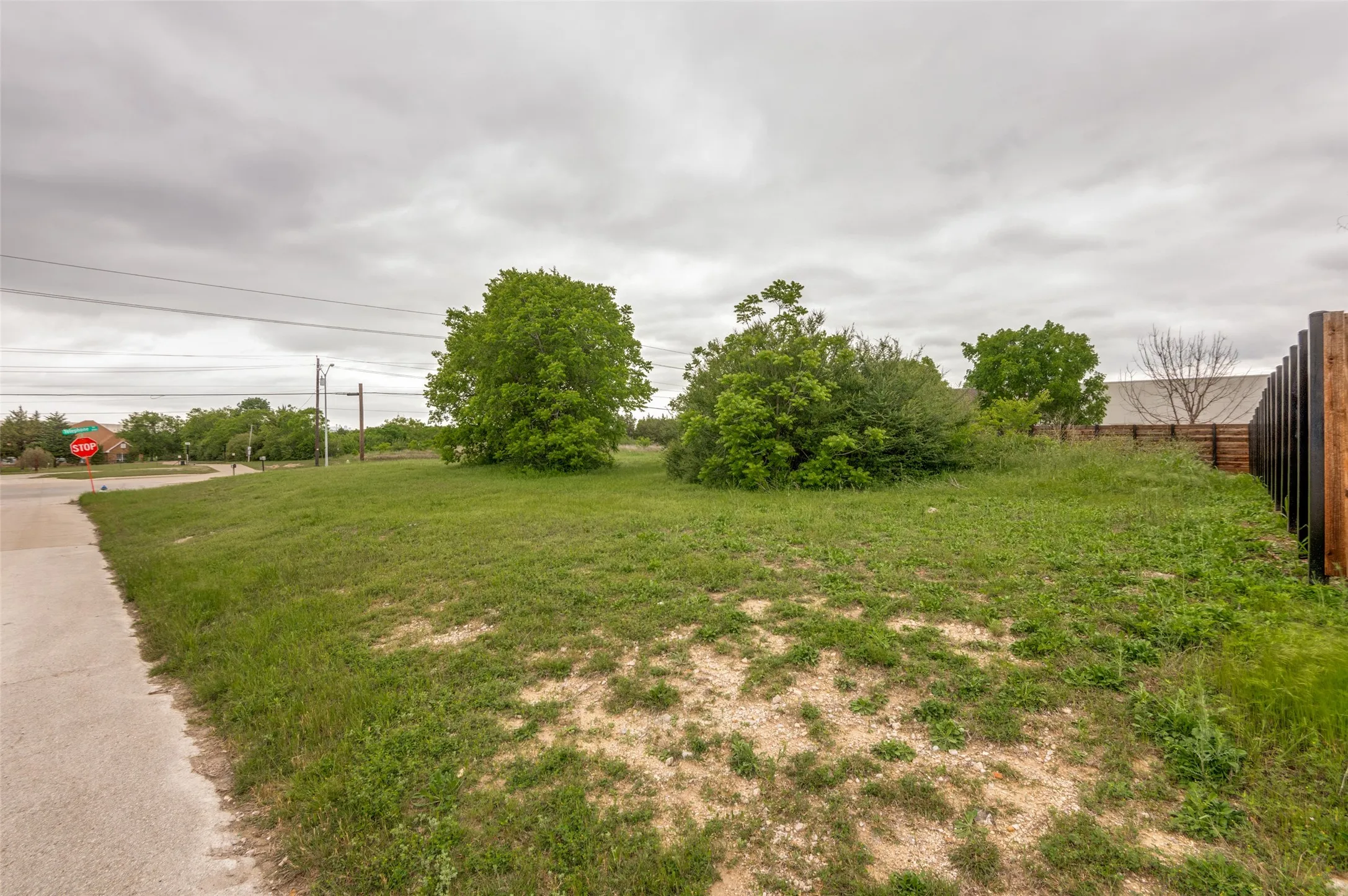 Expansive corner lot featuring mature trees, a paved curb, and an adjacent wood fence
