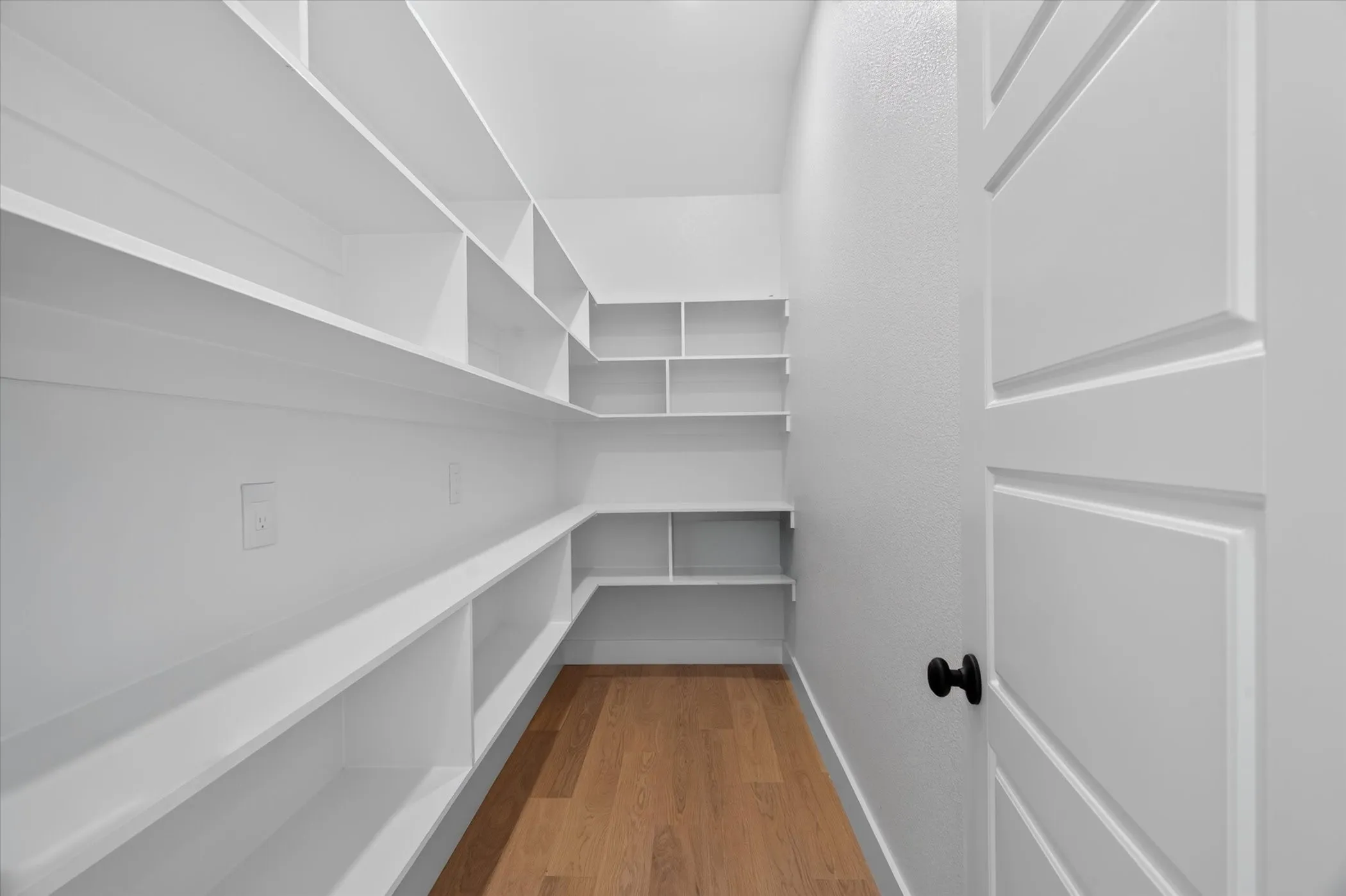 Expansive walk-in pantry with extensive built-in shelving, wood-finish flooring, and a paneled interior door with matte black hardware