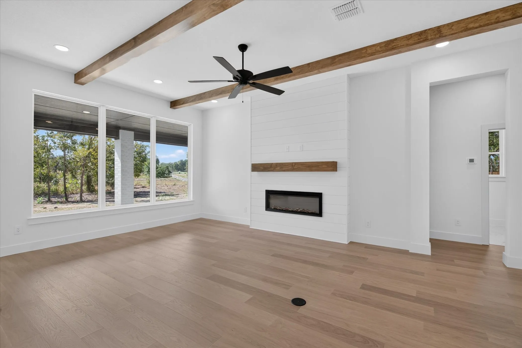 Expansive room featuring wood-finish flooring, exposed ceiling beams, an electric fireplace with a wood mantle, and large windows