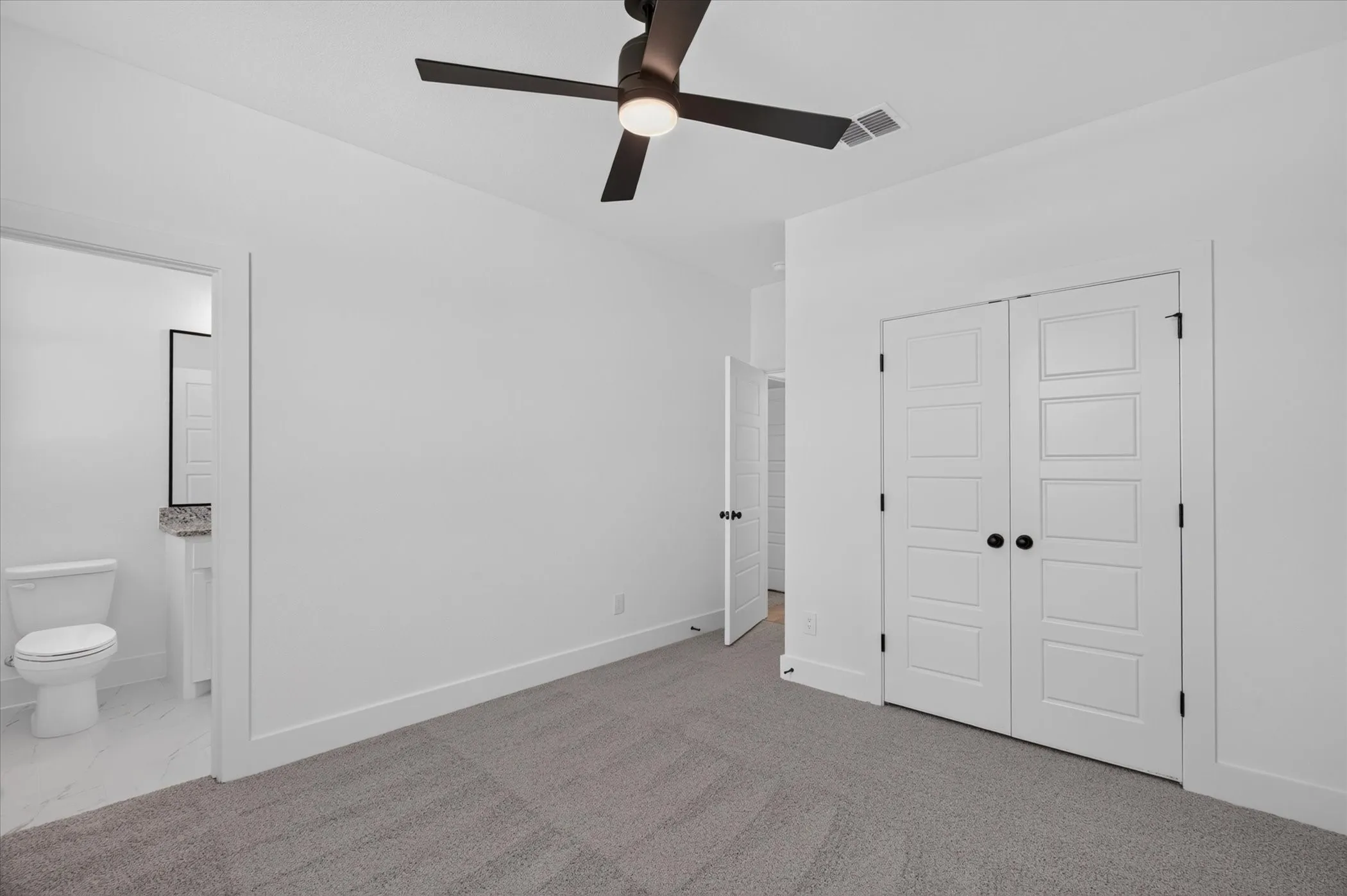 Spacious primary bedroom featuring carpeted flooring, a ceiling fan with integrated lighting, dual closets with six-panel doors, and an ensuite bathroom with toilet and shower enclosure