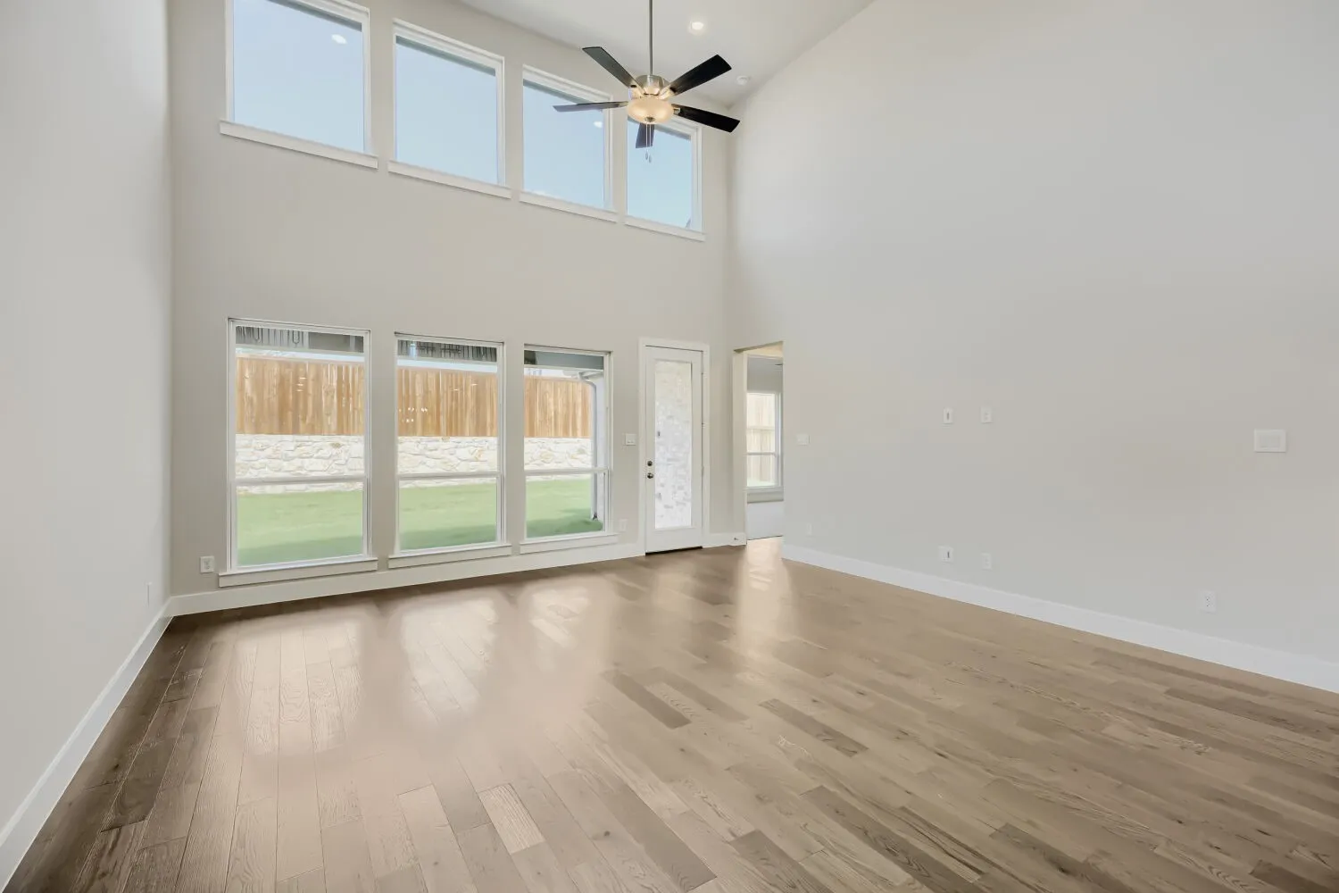 Expansive living space featuring soaring ceilings, wood-finish flooring, a contemporary ceiling fan, and multiple windows providing views of the outdoor area with a wood fence and stone accent wall
